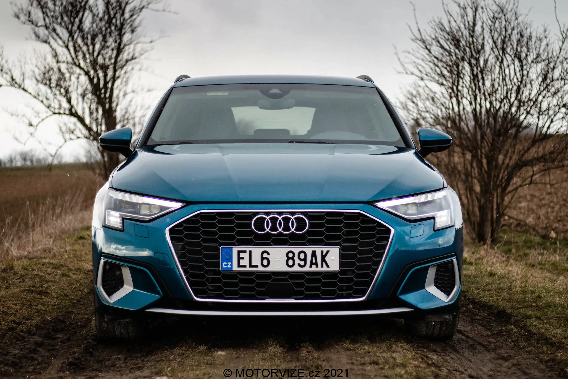 Predný pohľad na Audi A3 Sportback 2020 fotografovaný z nízkeho uhla. Obrázok sa zameriava na prednú časť auta, zobrazujúci osmihrannú mriežku Singleframe s včelími vzormi, ostré uhlové LED svetlá s výraznými dennými svetlami a veľké vzduchové vstupy vo tvarovanom prednom nárazníku. Štyri kruhy Audi sú namontované uprostred masky, s európskou registračnou značkou pod ním. Auto, jasné modré farby, je zaparkované na trávnatej ceste s oblačným nebom v pozadí, čo naznačuje jesenné alebo zimné obdobie.