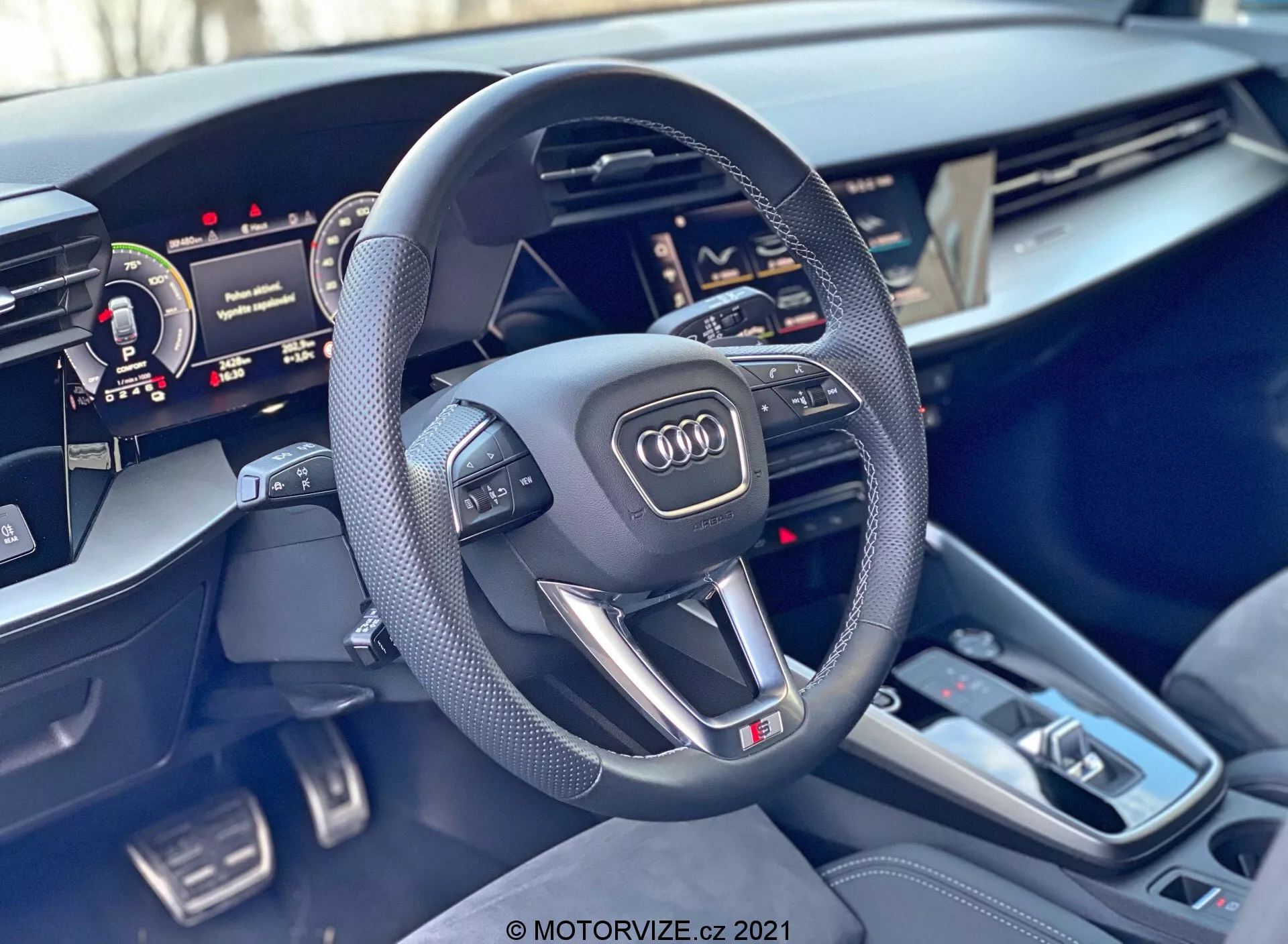 Pohľad do interiéru Audi A3 Sportback 2020 so zameraním na volant a stredovú konzolu. Športový trojramenný volant so splošteným spodkom má perforované kožené úchyty, multifunkčné tlačidlá, emblém Audi a značku 'S'. Za volantom je čiastočne viditeľný digitálny prístrojový panel a napravo čiastočne obrazovka Centrálnej multimediálnej rozhranie (MMI). Stredová konzola obsahuje radiacu páku automatickej prevodovky, niekoľko tlačidiel, dotykovú plochu, dva držiaky na nápoje a malé úložné priehradky. Interiérové materiály majú prémiové akcenty kartáčovaného kovu a kvalitné vyhotovenie, čo zdôrazňuje luxus a pokročilú technológiu vozidla.