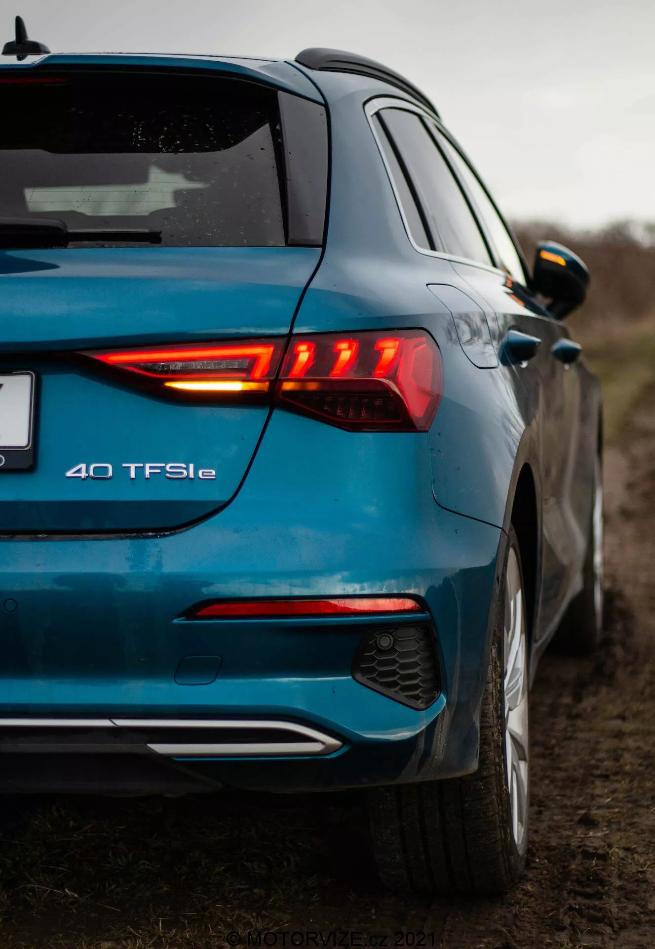 Pohľad z nízkeho uhlu na moderný modrý Audi A3 2020 (A3 Sportback) odfotený zo zadného trojštvrťového uhlu, zdôrazňujúci osvetlený zadný LED svetelný klaster, označenie modelu 40 TFSIe, dvojfarebný zadný nárazník so zabudovaným difúzorom a parkovacími senzormi, kovový lak, strešný spojler, farebne zladené bočné zrkadlá s integrovanými smerovkami a náznak predných dverí a bočného profilu. Fotografia zdôrazňuje dizajnové prvky a značenie vozidla v vidieckom prostredí s oblačným nebom a nízkou hĺbkou ostrosti.