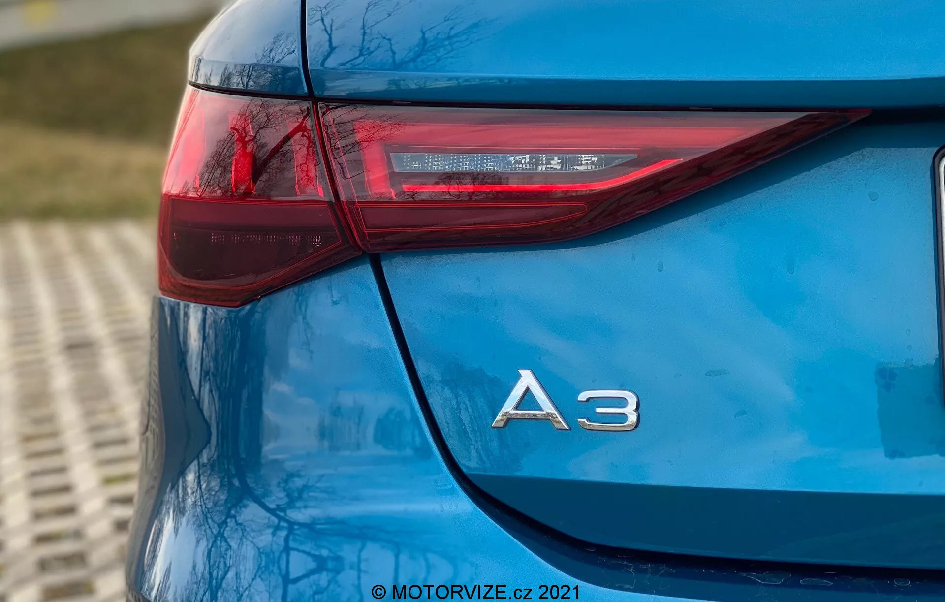 Tento obrázok zobrazuje detailný nízkouhlový záber na ľavý zadný roh modrej Audi A3 Sportback 2020. Fotografia zdôrazňuje elegantný dizajn zadného svetla, ktoré má ostrú horizontálnu orientáciu zasahujúcu do boku vozidla, a chrómové označenie modelu 'A3' tesne pod ním. LED prvky v zadnom svetle zdôrazňujú pokročilú osvetľovaciu technológiu vozidla. Lesklý lak a celková čistota modelu ukazujú vysokokvalitné ošetrenie vozidla, pričom mierne rozmazané pozadie naznačuje vonkajšie prostredie. Vodotlač '© MOTORVIZE.cz 2021' označuje vlastníctvo a pôvod obrázku.