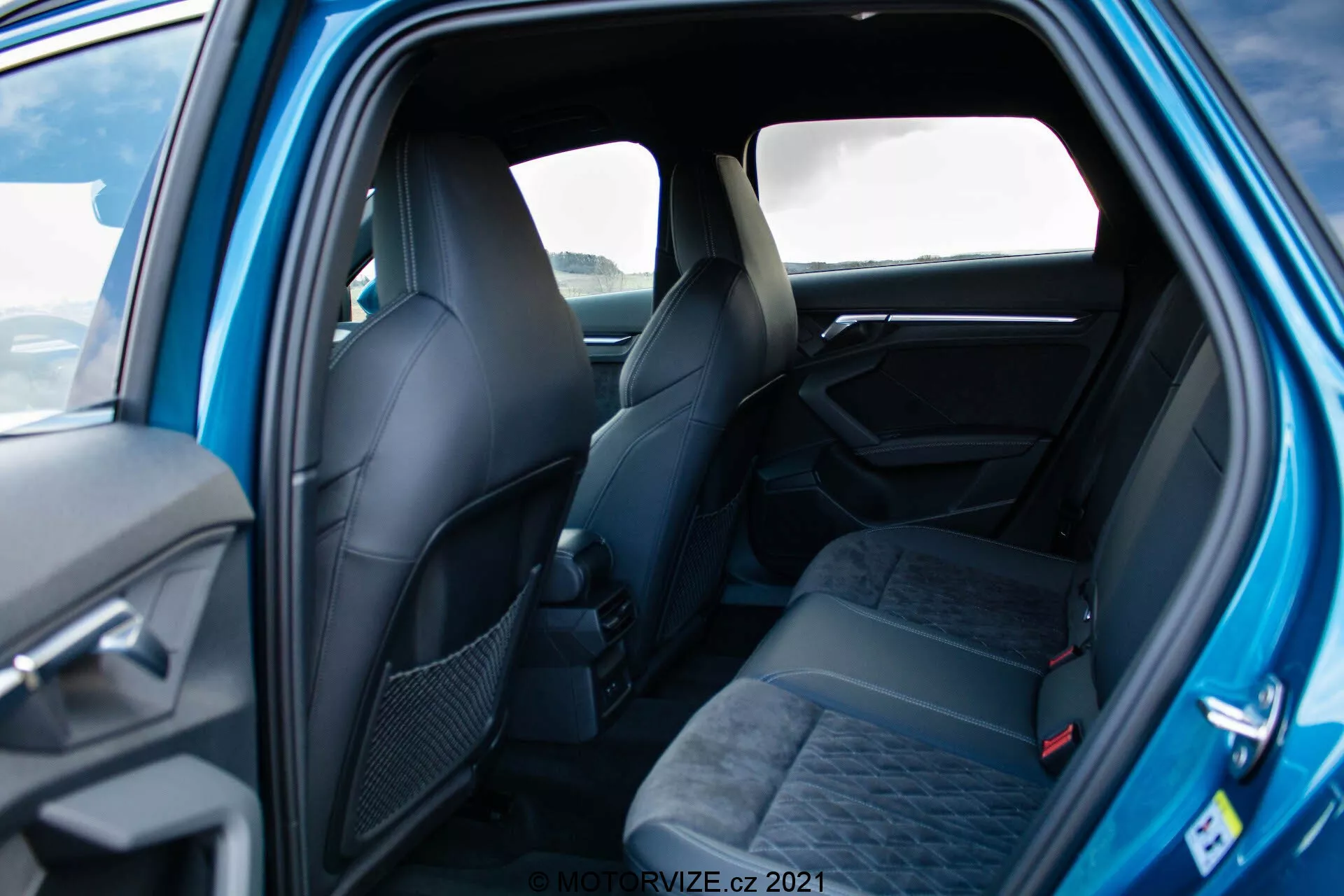 Záber interiéru vozidla Audi A3 Sportback 2020 z pohľadu zo zadných dverí ukazuje priestranné sedadlá pre troch pasažierov s opierkami hlavy a bezpečnostnými pásmi, čalúnené tmavou látkou a vzorované. Čiastočne viditeľné sú predné sedadlá s ergonomickým dizajnom. Viditeľné prvky dverného panelu zahŕňajú rukoväť a možné ovládacie prvky. Dobre osvetlený záber zdôrazňuje elegantný, prémiový pocit s kvalitnými materiálmi a dizajnovými detailmi.