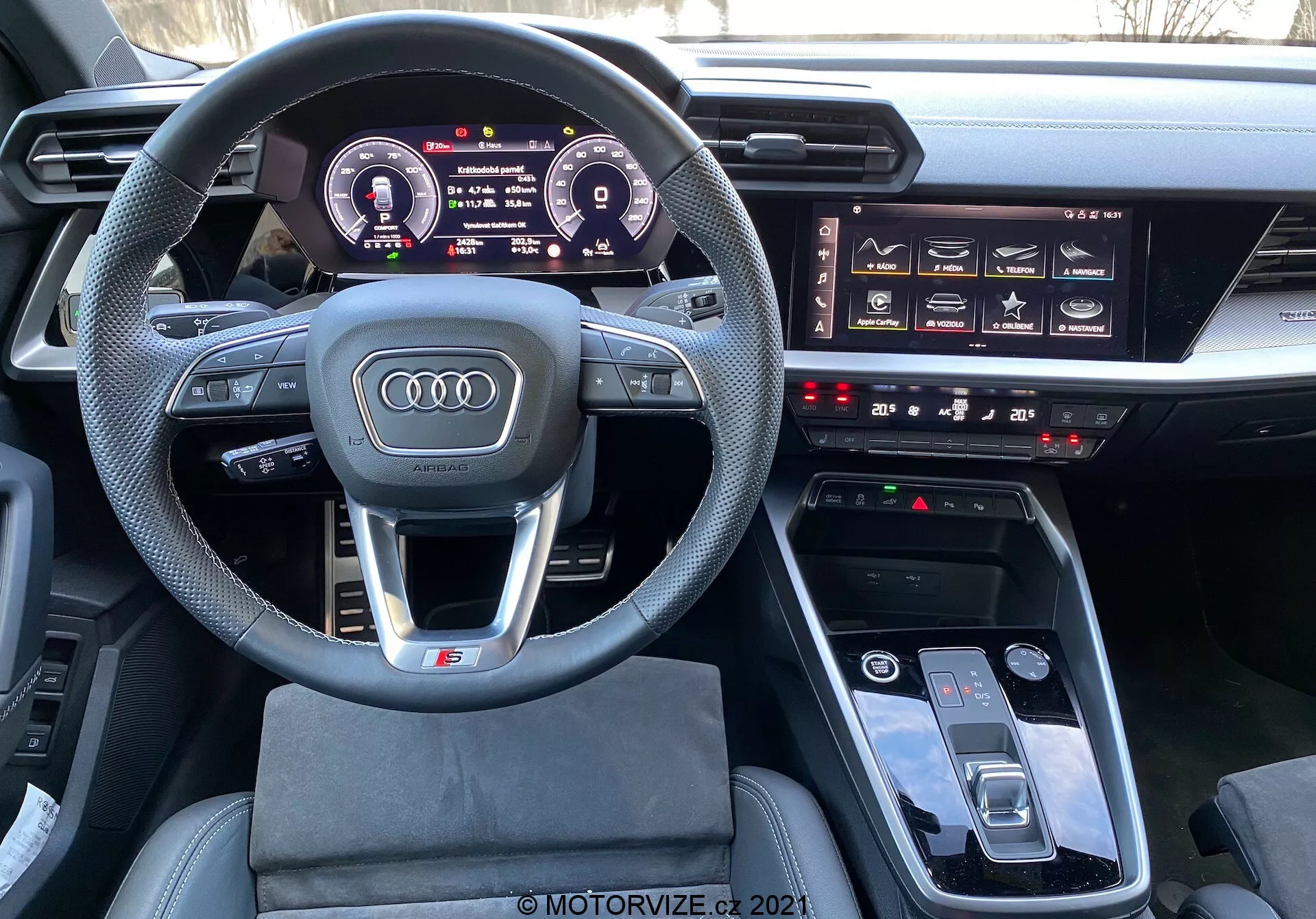 Pohľad z vodičovho sedadla na interiér Audi A3 Sportback 2020, zobrazujúci multifunkčný športový volant obalený kožou s plochým spodkom, digitálny prístrojový panel Audi Virtual Cockpit, významný infotainment systém, hmatové tlačidlá na ovládanie klimatizácie a dotykovú podložku MMI v stredovej konzole, naznačujúci moderný, luxusný a technologicky pokročilý interiér automobilu.