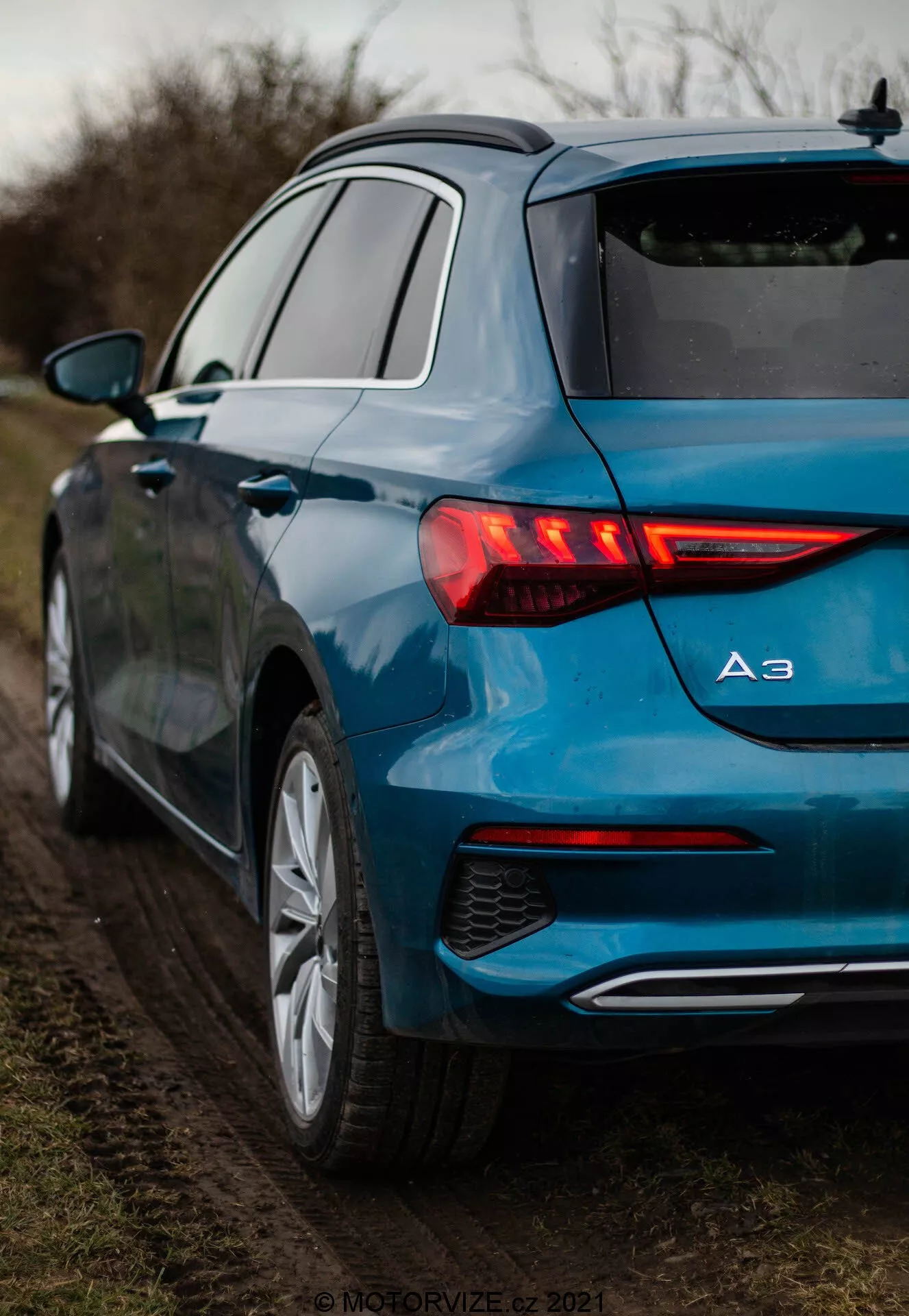 Detailný pohľad na metalicky modrú Audi A3 2020 Sportback zo zadnej trojštvrťovej perspektívy, zdôrazňujúci jej osvetlené LED zadné svetlá s dynamickými smerovkami, modelový znak zobrazujúci 'A3', viacramenné zliatinové kolesá a jeden výfuk. Auto je zaparkované na nespevnenej krajnici obklopenej vegetáciou, pri mäkkom, rozptýlenom osvetlení.