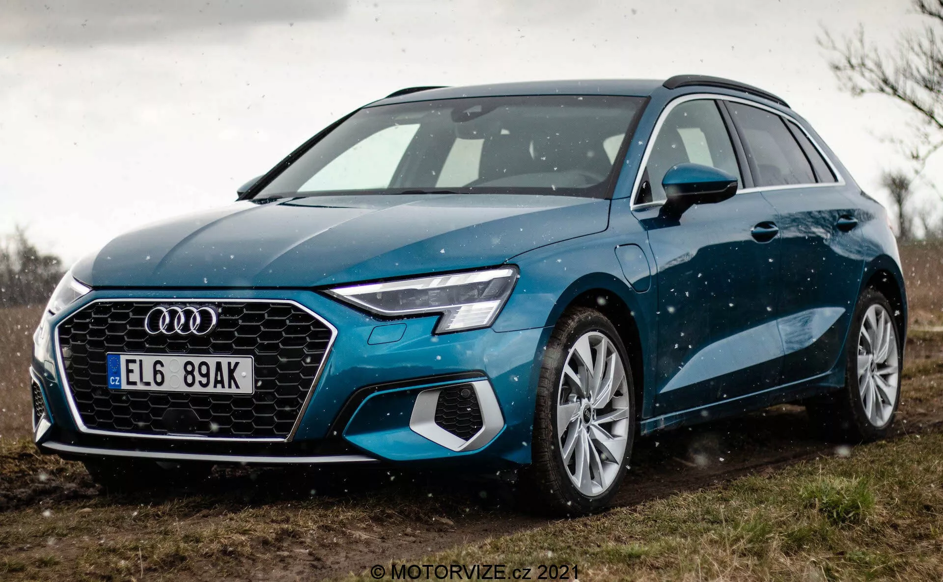 Nízkoúhlový pohľad na modrý Audi A3 Sportback 2020, zvýrazňujúci prednú masku a dizajn LED svetlometov. Blízky záber sa sústreďuje na prednú ľavú stranu vozidla, ukazujúc jeho šesťhrannú masku s výrazným logom Audi, integrovanými senzormi pre funkcie ADAS, veľkými kolesami z ľahkej zliatiny s viacerými lúčmi a elegantnými bočnými zrkadlami. Auto je zaparkované na nespevnenom povrchu so zeleňou v pozadí, čo naznačuje vidiecke prostredie. Jemné kvapôčky vody na karosérii vozidla zvýrazňujú jeho línie a farbu.