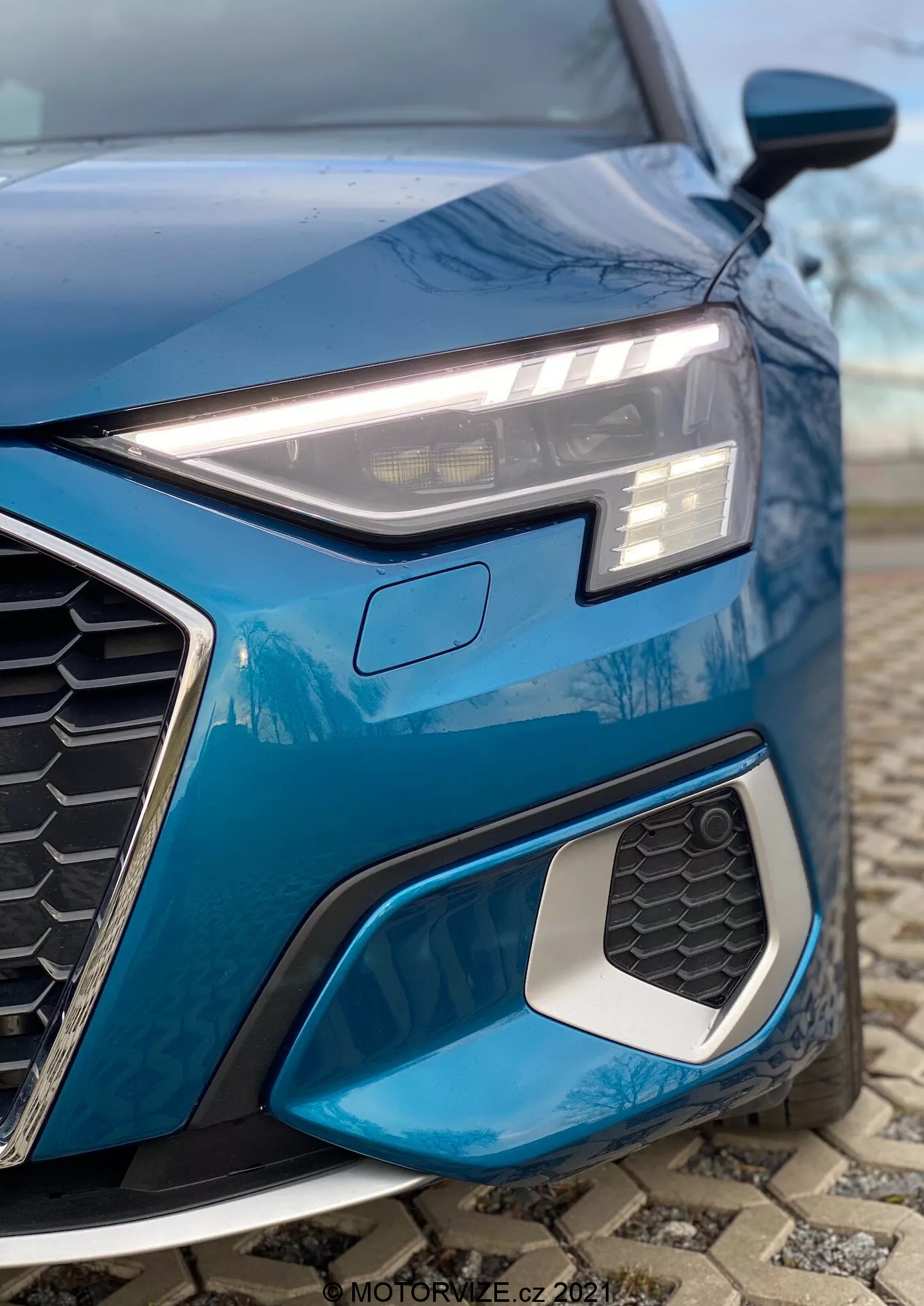Detailný záber na ľavú prednú štvrtinu modrého Audi A3 2020 (A3 Sportback), zdôrazňujúci elegantný, uhlový LED svetlomet so svetlami denného svietenia, výraznú šesťuholníkovú masku chladiča s motívom včelích plastov a prívod vzduchu s kovovou hranou. Záber z nízkeho uhla podčiarkuje športové dizajnové prvky, zložitý geometrický dizajn hmlovky a rozmazané pozadie pre zameranie sa na detaily.