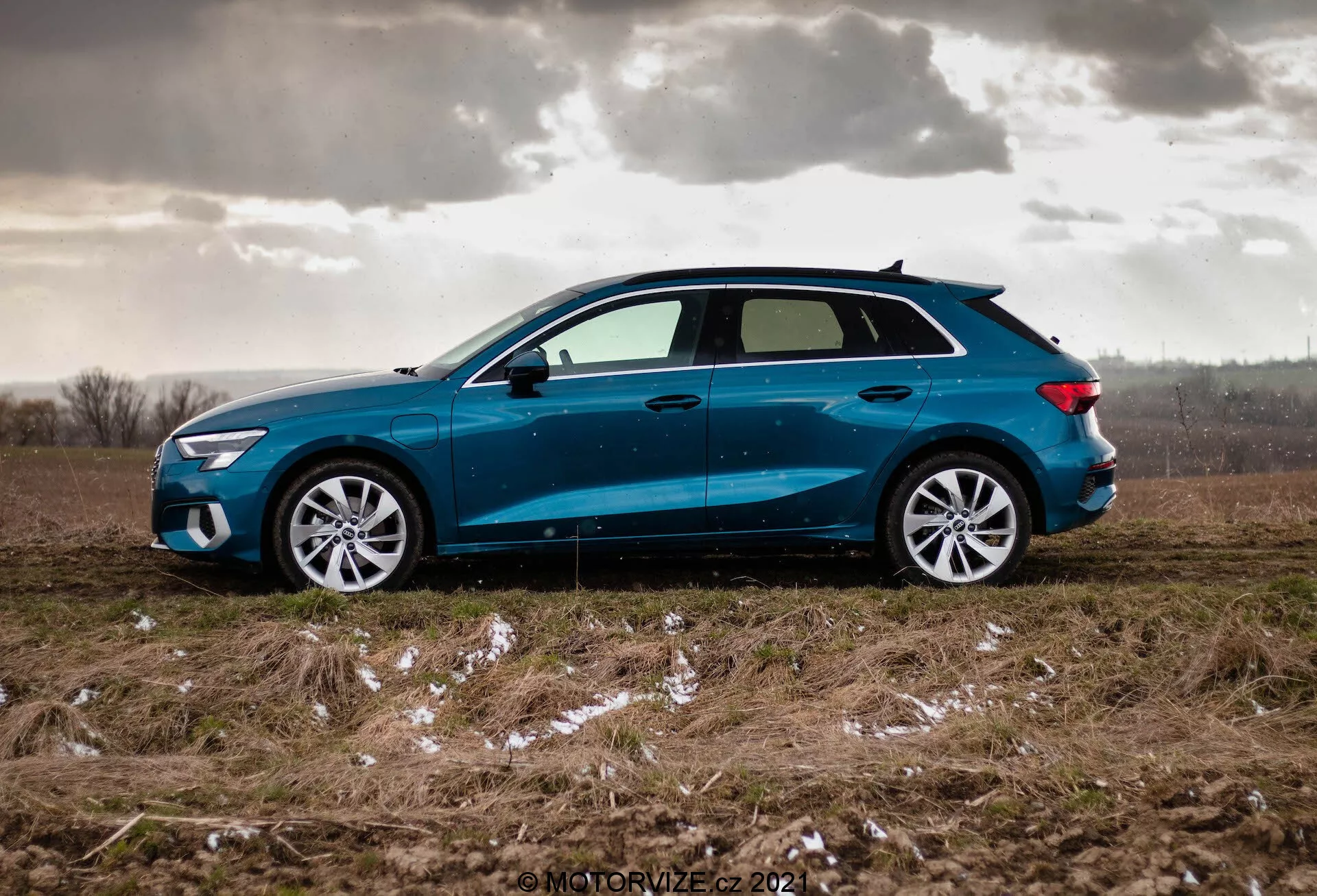 Bočný profil modrého Audi A3 Sportback z roku 2020 zaparkovaného na nerovnom trávnatom poli s náznakmi snehu alebo ľadu a zatiahnutou oblohou, zdôrazňujúci dynamický dizajn auta s mierne sklonenou strešnou líniou, viaclúčovými zliatinovými kolesami, mierne zatienenými oknami, bočnými spätnými zrkadlami vo farbe karosérie s integrovanými blikačmi a ostrými LED svetlometmi a zadnými svetlami.