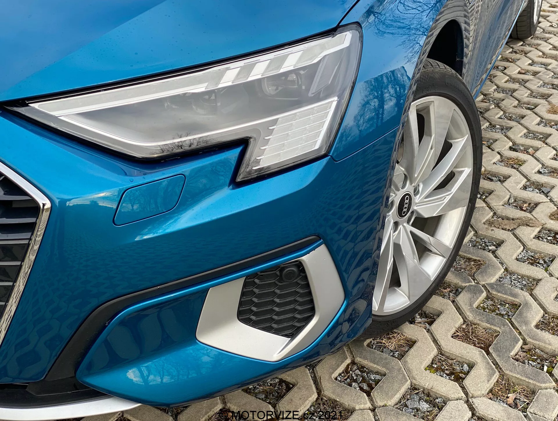 Detailný pohľad na prednú ľavú štvrtinu kovovo modrého Audi A3 2020 Sportback. Obrázok, urobený z nízkeho uhla, zdôrazňuje kľúčové dizajnové prvky, vrátane moderného LED svetlometu s individuálnymi svetelnými prvkami, viacramenného zliatinového kolesa, uhlového predného nárazníka s výraznou voštinovou mriežkou a dlažby z dlažobných kociek v mestskom prostredí.