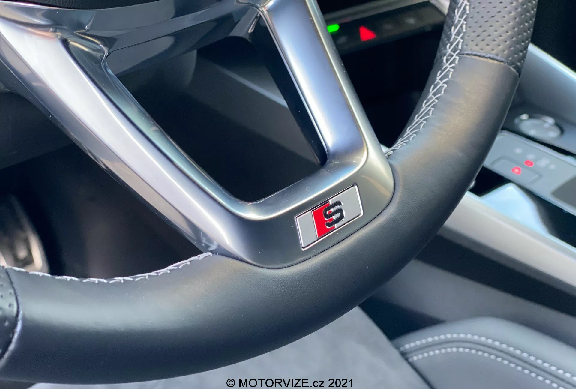 Detailný pohľad na volant moderného auta z pohľadu vodiča, zameraný na spodnú časť s výrazným odznakom 'S' olemovaným červenou. Volant zobrazuje kombináciu hladkej kože a lesklejšieho materiálu s viditeľným šitím, zdôrazňujúcim remeselné spracovanie a športový charakter Audi A3 2020 Sportback.