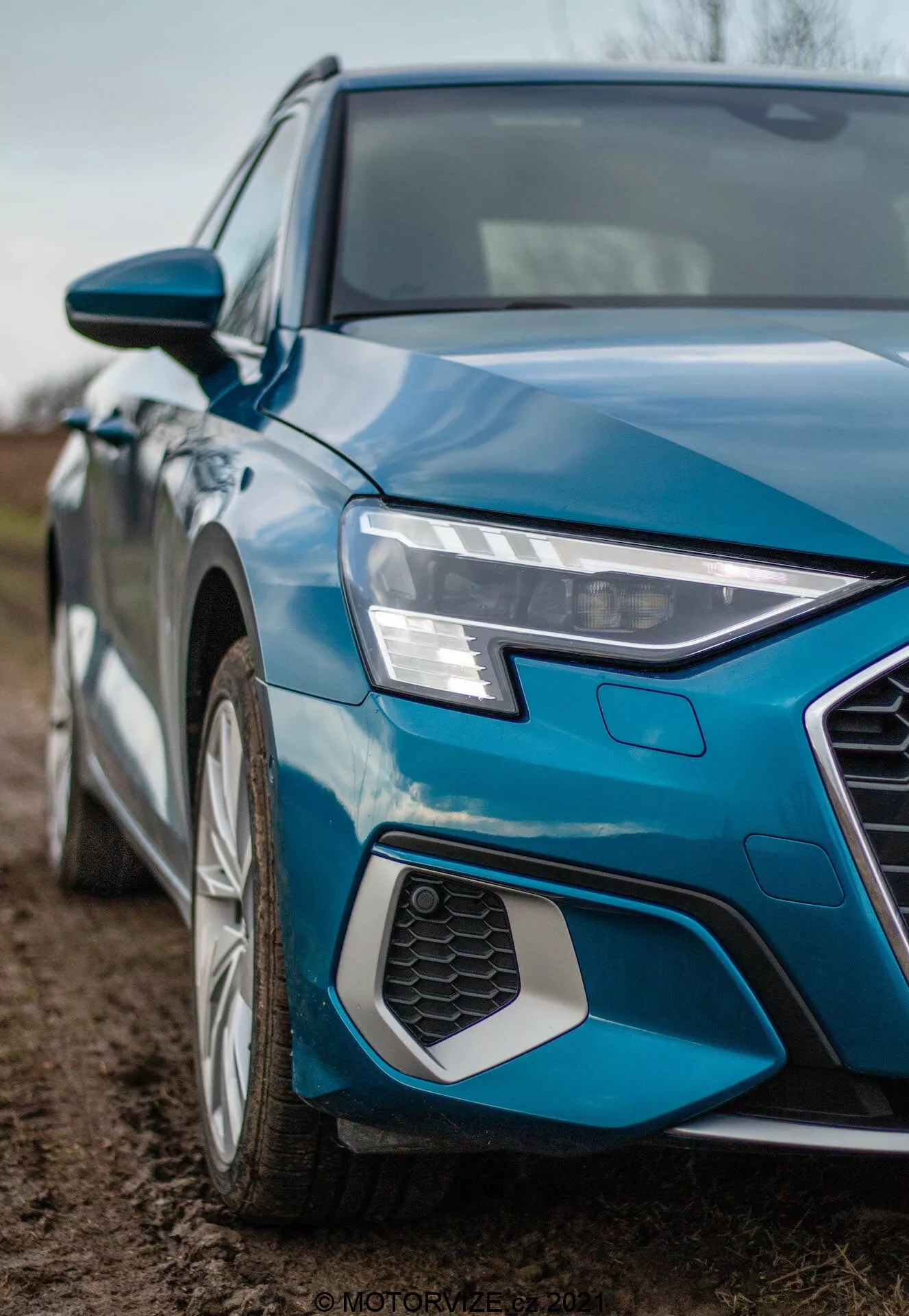 Detailný záber Audi A3 Sportback 2020 z šikmého uhla, zameraný na ľavý predný kvadrant. Fotografia z nízkeho uhla zachytáva ostrý a rohový predný ľavý svetlomet s LED denným svietením, časť šesťuholníkovej mriežky s medovým vzorom, ľavý predný blatník a segment dverí vodiča. Vidieť tiež viacramenné hliníkové koleso, kovovo modrý lak a spätné zrkadlo vo farbe karosérie s integrovanými smerovými svetlami, zdôrazňujúce moderný dizajn a technologické prvky.