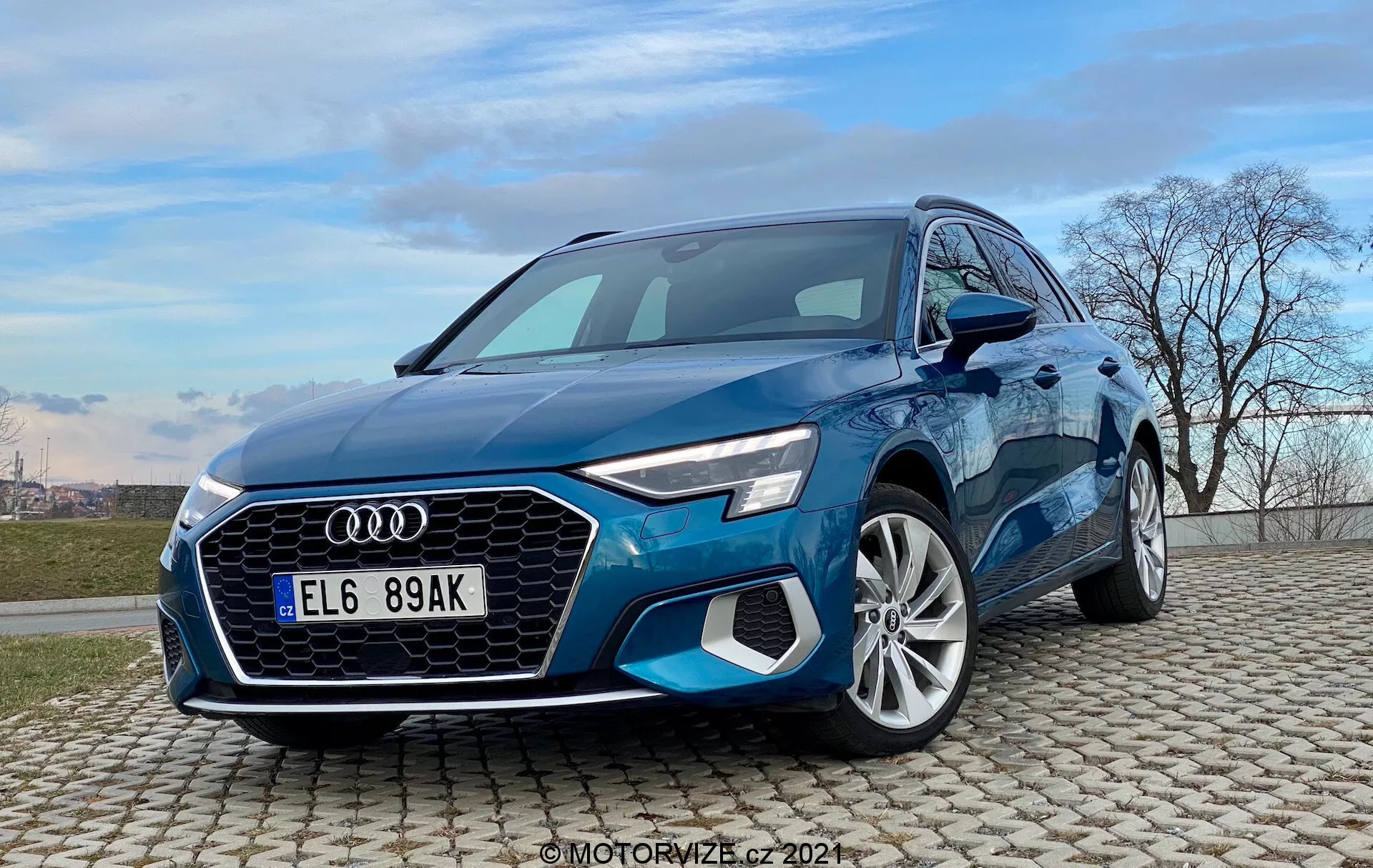 Trojštvrťový pohľad na modrý Audi A3 Sportback 2020 zachytený z nízkeho uhla. Obrázok zobrazuje dynamický a impozantný vzhľad auta s charakteristickou maskou Singleframe, šesťhranným tvarom s vertikálnymi lamelami, ostrými LED svetlometmi a vzduchovými vstupmi na prednom nárazníku. Sochársky tvarovaná kapota vedie k čelnému sklu, odhaľujúc aerodynamické kontúry a línie karosérie. Bočný profil zdôrazňuje definované blatníky nad viacramennými zliatinovými kolesami a šikmé A-stĺpiky s elegantnými bočnými zrkadlami. Auto je umiestnené na dlažobných kockách pod jasným modrým nebom s oblakmi a holou líniou stromov, čo naznačuje propagačný alebo recenzný kontext.