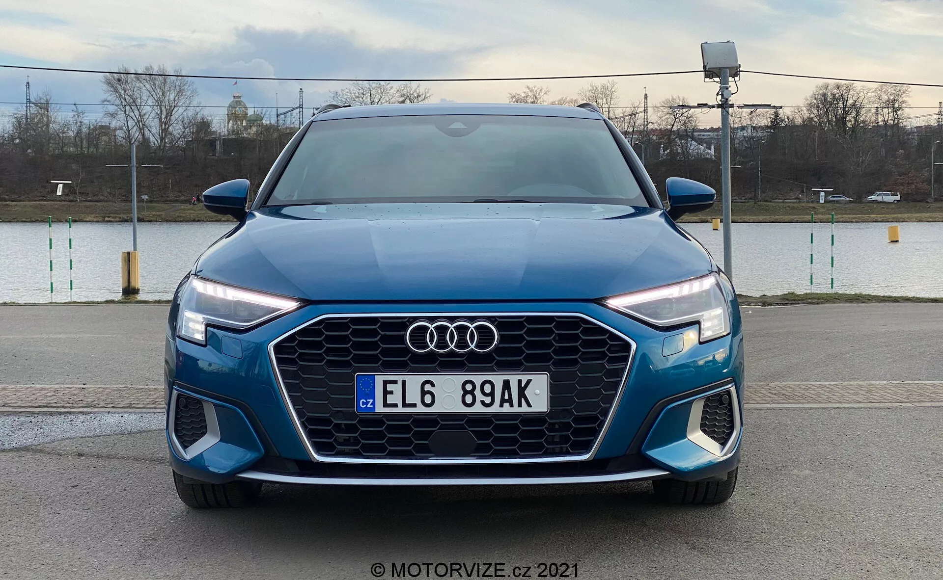 Čelný pohľad na modrý Audi A3 Sportback 2020, ktorý sa pýši výraznou maskou Singleframe s hexagonálnym dizajnom, logom Audi uprostred, LED svetlomety s denným svietením a predným nárazníkom s ďalšími vzduchovými otvormi a možnými hmlovými svetlami, na pozadí vonkajšieho prostredia.