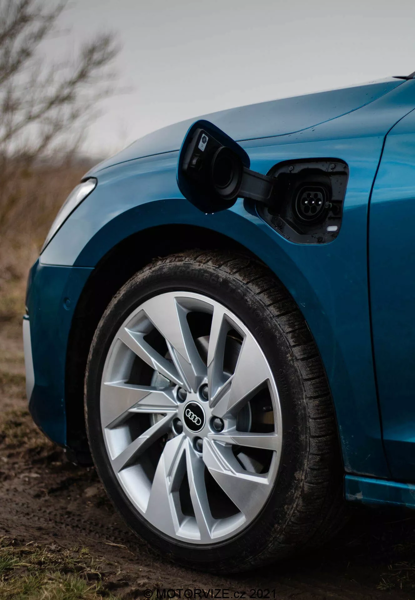 Detailný záber na prednú časť vodičovej strany modrého Audi A3 2020 Sportback, zdôrazňujúci koleso a nabíjací port. Obrázok, urobený z nízkeho uhla smerom dopredu, zobrazuje otvorený nabíjací port nad predným kolesom s pripojeným nabíjacím káblom. Plytká hĺbka ostrosti ostro zaostruje na koleso a nabíjací port, odhaľujúc typické Audi logo prelínajúcich sa kruhov a viaclúčový dizajn kolesa s nízkoprofilovou pneumatikou, všetko v mierne tlmenom, možno súmrakovom svetle.