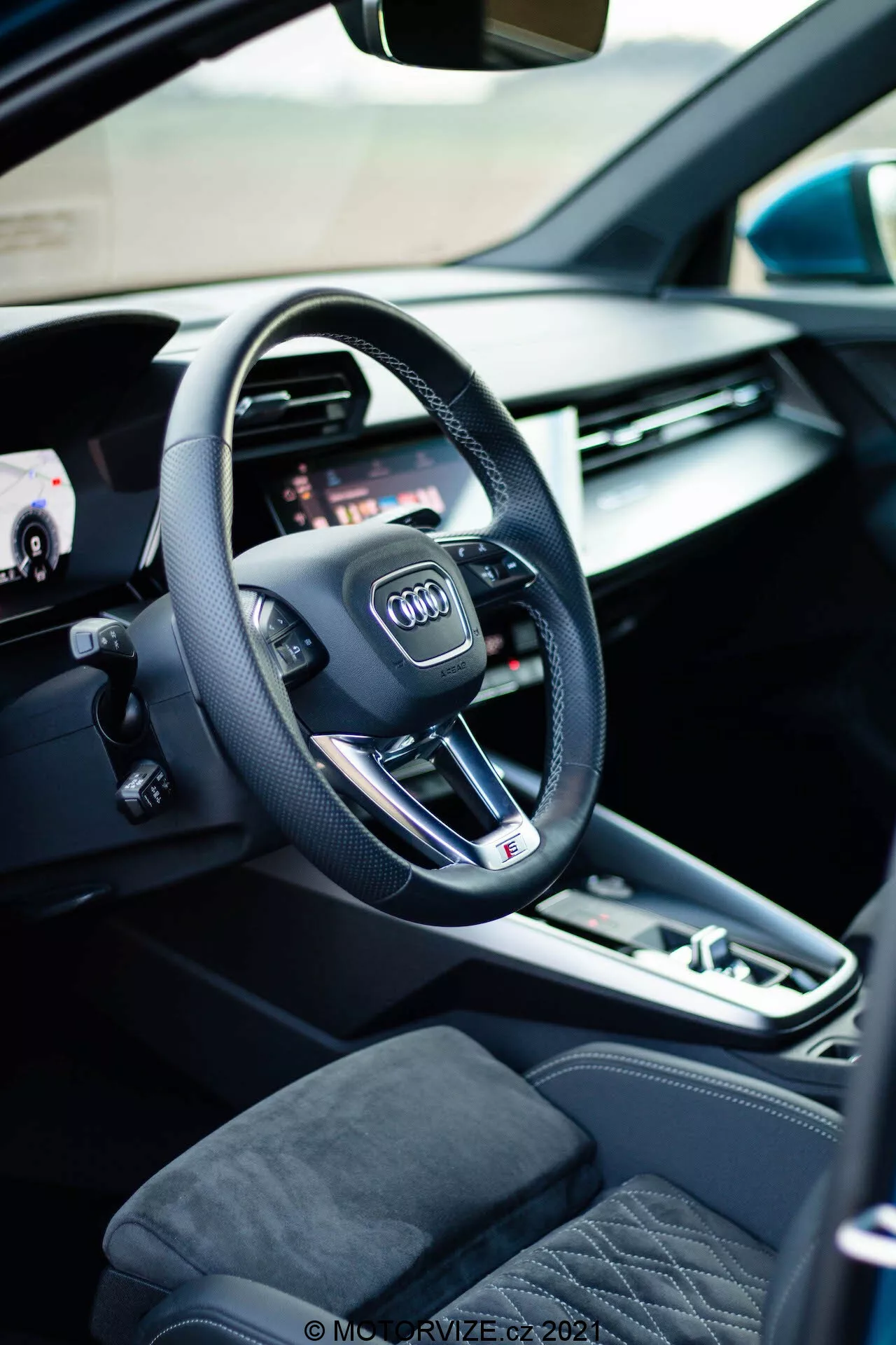Interiérový pohľad na Audi A3 Sportback 2020, zameraný na stranu vodiča s moderným trojramenným volantom s logom Audi, integrovanými multifunkčnými tlačidlami a plochým spodným dizajnom, ktorý naznačuje športový výkon. Za volantom je viditeľná časť digitálneho prístrojového panela alebo virtuálneho kokpitu, ktorý ponúka prispôsobiteľné možnosti zobrazenia pre rôzne funkcie auta. Naľavo, otvorené dvere od auta odhaľujú ovládanie elektrických okien a pamäťové nastavenia sedadiel, zdôrazňujúc pohodlie a prispôsobenie. Stredová konzola zahŕňa automatický volič prevodovky, tlačidlo elektronickej parkovacej brzdy a dotykovú podložku/ovládacie koliesko pre infotainment systém, zdôrazňujúc zapojenie vodiča. Viditeľné sedadlá sú čalúnené prémiovými materiálmi s prešívaným dizajnom, prispievajúcim k pocitu luxusu v interiéri.