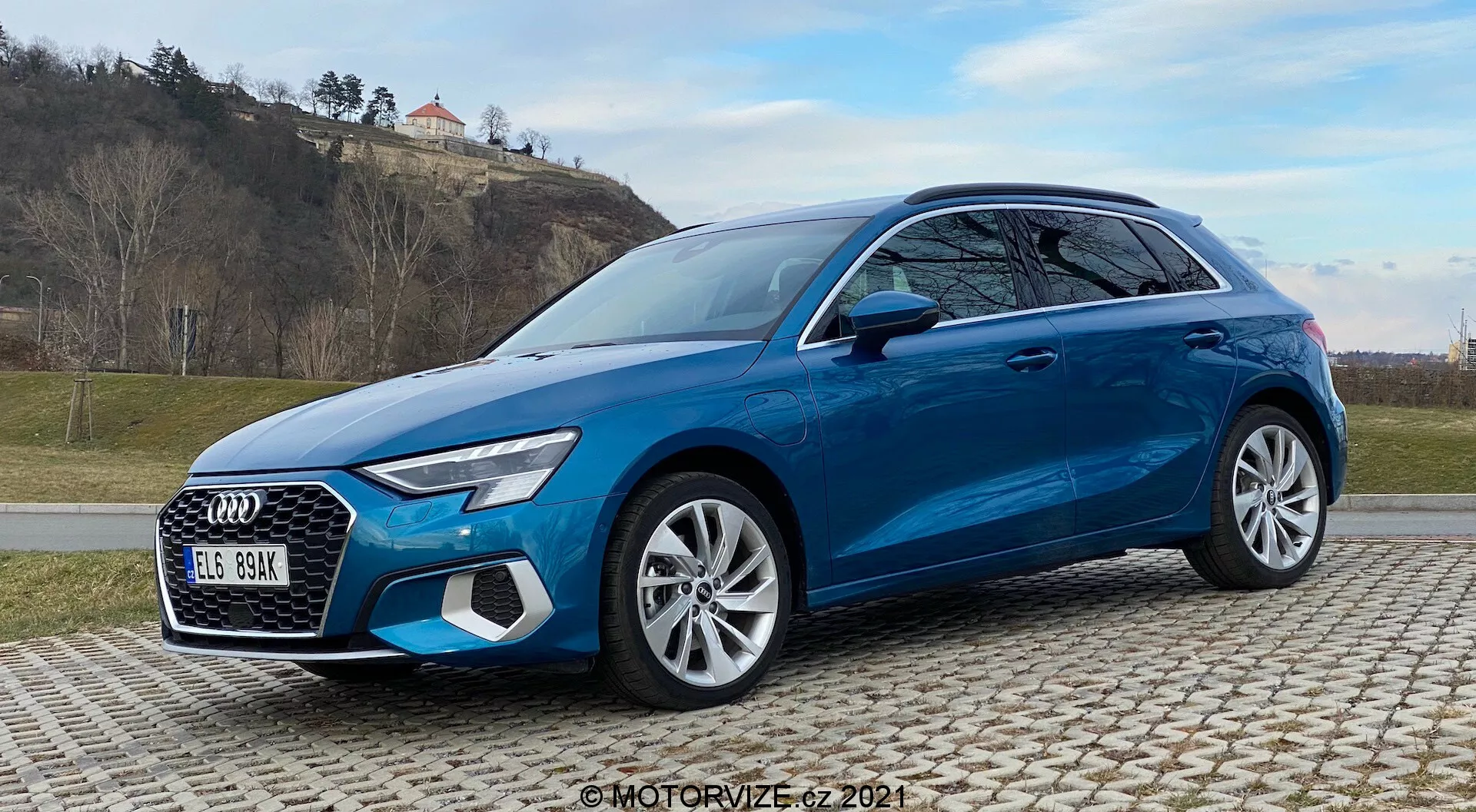 Pohľad z predu pod uhlom tri štvrtiny na svetlomodré Audi A3 Sportback 2020 na dláždenej ploche, ukazujúci výrazné dizajnové prvky ako LED svetlomety, výrazná mriežka, viacramenné zliatinové kolesá a pokročilé bezpečnostné systémy. Zachytené s trávnatým, kopcovitým pozadím a čiastočne zatiahnutou oblohou.