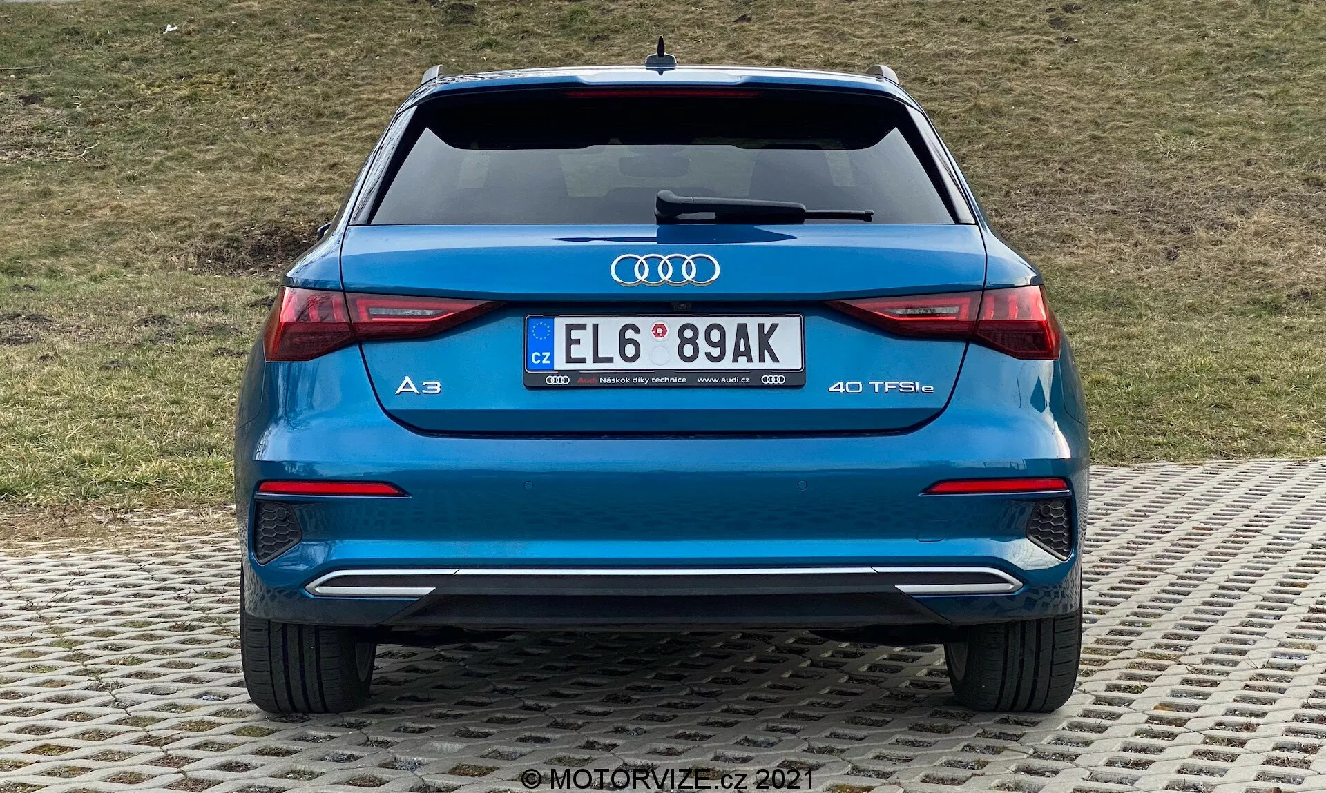 Zadný pohľad na modrý Audi A3 2020 Sportback, ktorý ukazuje zadný nárazník, kufor, zadné svetlá, výfuky a zadné sklo. Výrazné logo Audi a označenie modelu 'A3' s odznakom '40 TFSI' naznačujúci turbomotor na benzín. LED zadné svetlá s elegantným horizontálnym dizajnom, dvojité výfuky, zadný nárazník s difúzorom a parkovacími senzormi a európska ŠPZ. V pozadí trávnatý svah.