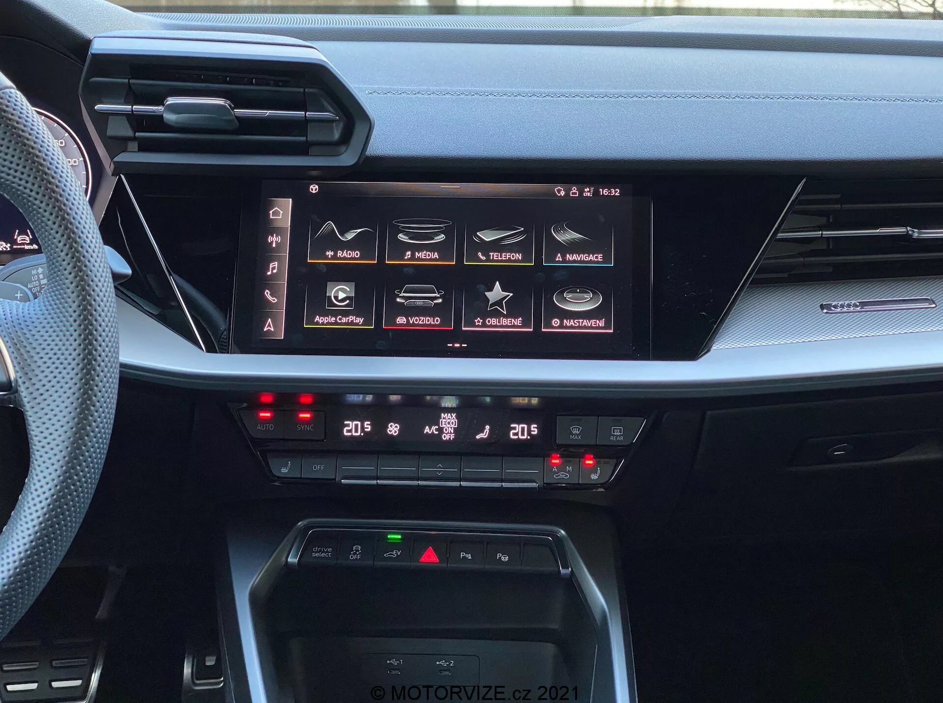 Pohľad na interiér palubnej dosky modelu Audi A3 Sportback 2020, zdôrazňujúci jeho pokročilý infotainment systém a ovládanie klimatizácie. Obrázok zachytáva veľký horizontálny dotykový displej s funkciami ako navigácia, médiá, telefonické pripojenie a nastavenia vozidla, naznačujúci komplexné schopnosti infotainmentu vrátane Apple CarPlay a Android Auto. Pod displejom sú dvojzónové digitálne ovládače klimatizácie s nastavením na 20 stupňov Celzia pre vodiča aj spolujazdca. Palubná doska tiež obsahuje oblasť pre bezdrôtové nabíjanie a tenké horizontálne vetracie otvory, ktoré ilustrujú moderný dizajn automobilových interiérov. Fotografia je označená vodoznakom 'MOTORVIZE.CZ 2021'.