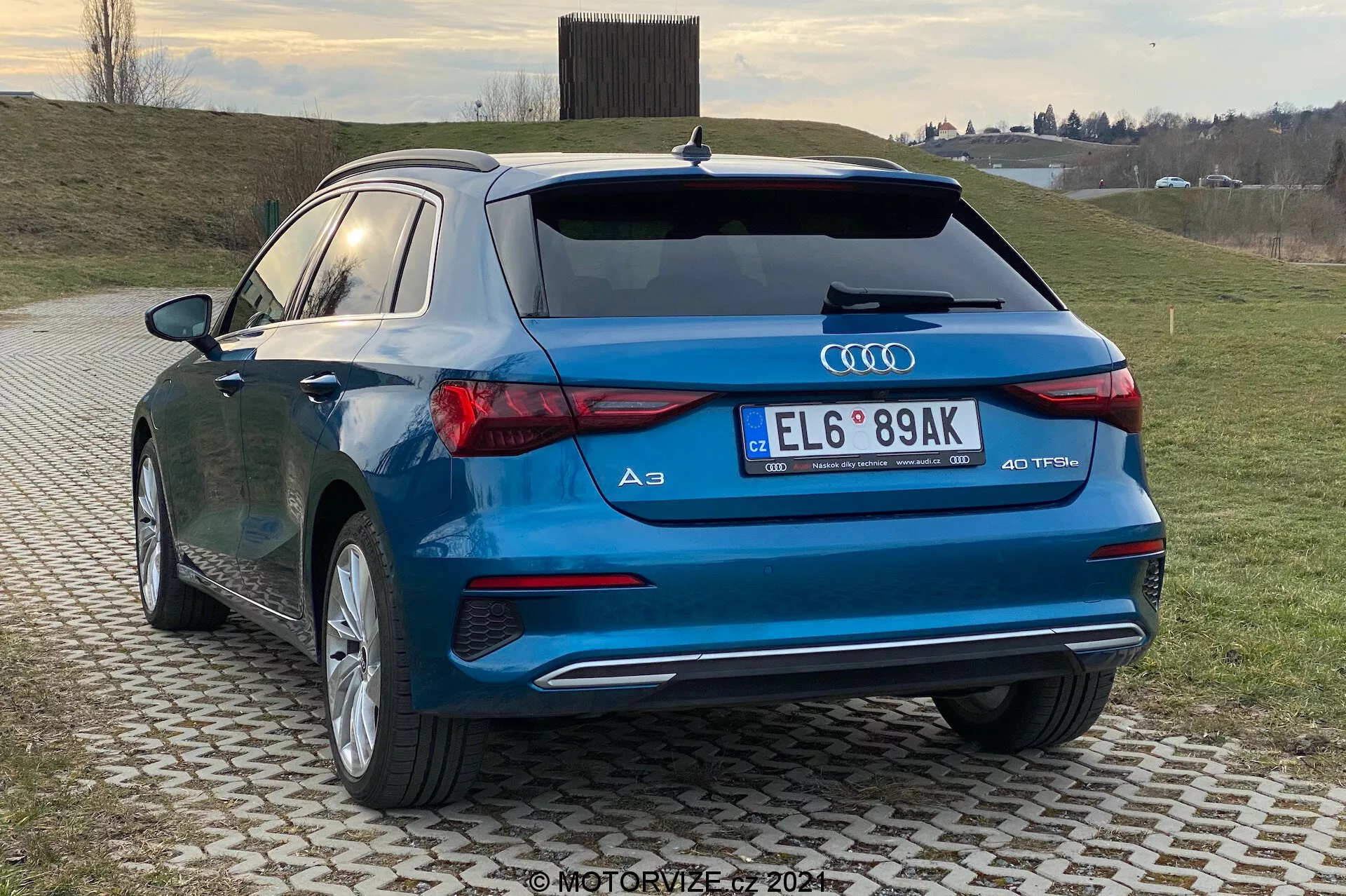 Obrázok zachytáva moderný hatchback, Audi A3 Sportback 2020, z pohľadu zadnej tretiny, zvýrazňujúc zadný a bočný profil vozidla. Charakteristické dizajnové prvky ako LED zadné svetlá, dvojité výfukové koncovky, emblém Audi kroužkov a značka '40 TFSI' sú viditeľné, čo naznačuje športovú variantu S-line. Auto s lesklým modrým lakom je zaparkované na vzorovanej dlažbe v vidieckom alebo polo-vidieckom prostredí s trávnatým pozadím a zatiahnutou oblohou.