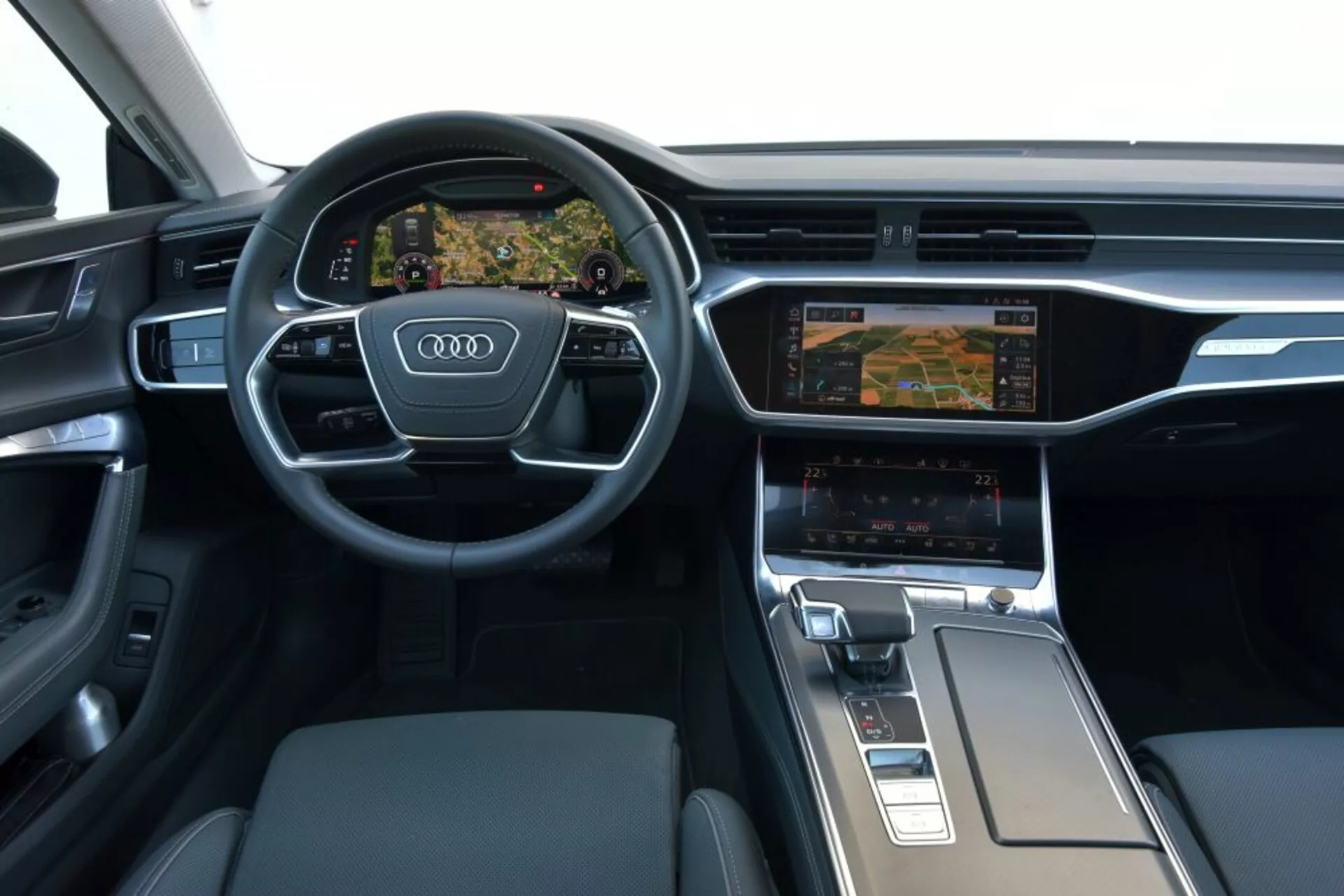 Interiér Audi A7 Sportback 2018 so zameraním na palubnú dosku a oblasť stredovej konzoly vodiča, predstavujúci trojramenný multifunkčný volant s logom Audi, digitálny prístrojový panel Virtual Cockpit zobrazujúci navigáciu, duálny displej infotainment systému s nastaveniami klimatizácie, ergonomickú radiacu páku a prémiové materiály ako brúsený kov a koža.