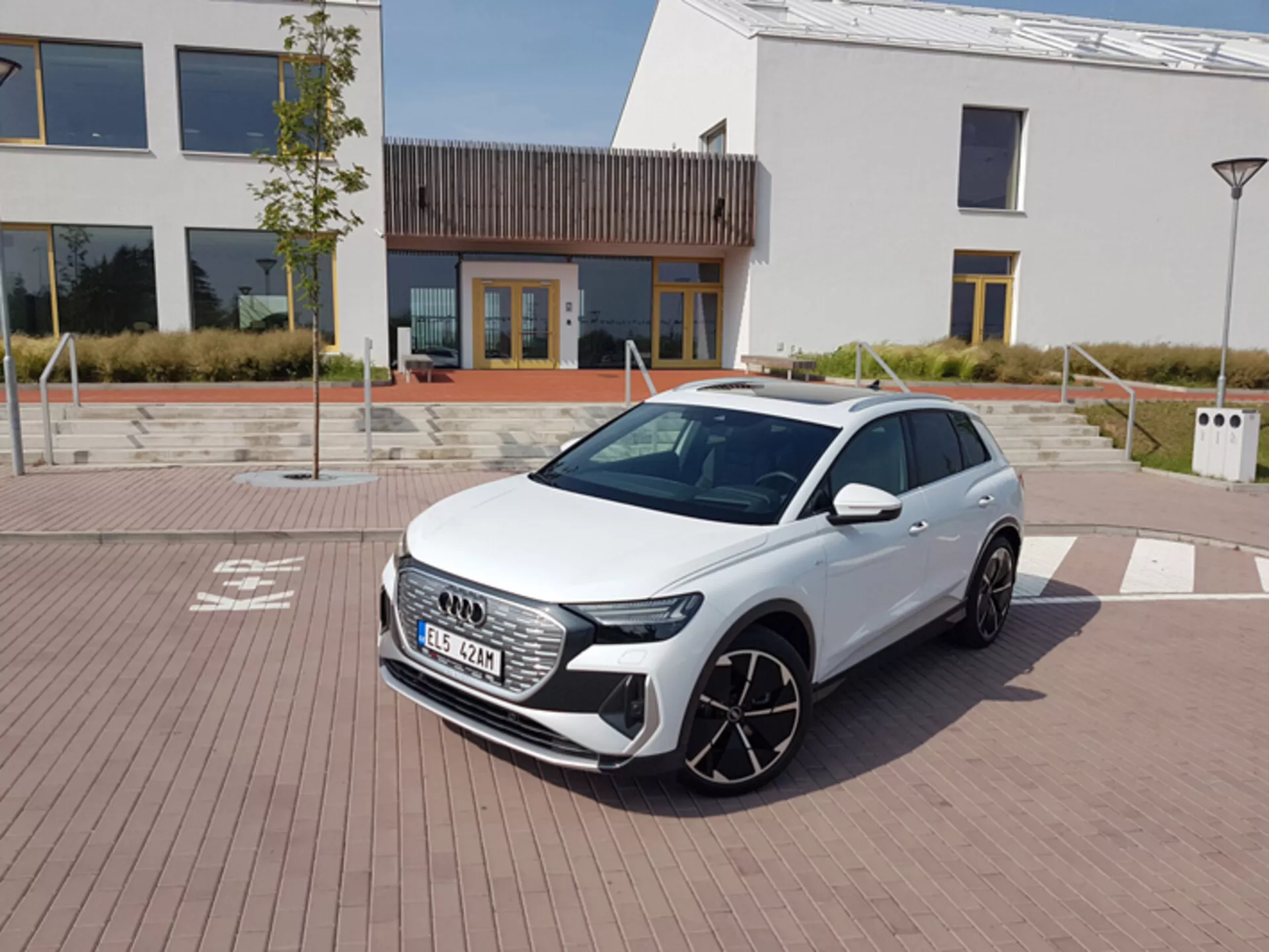 Biela Audi Q4 e-tron 2021 s modernými LED svetlometmi, šesťuholníkovou prednou mriežkou a veľkými zliatinovými kolesami zaparkovaná na dlažbe s tehlovým vzorom s mestskou architektúrou a nabíjacou stanicou na pozadí pod jasnou poludňovou oblohou.