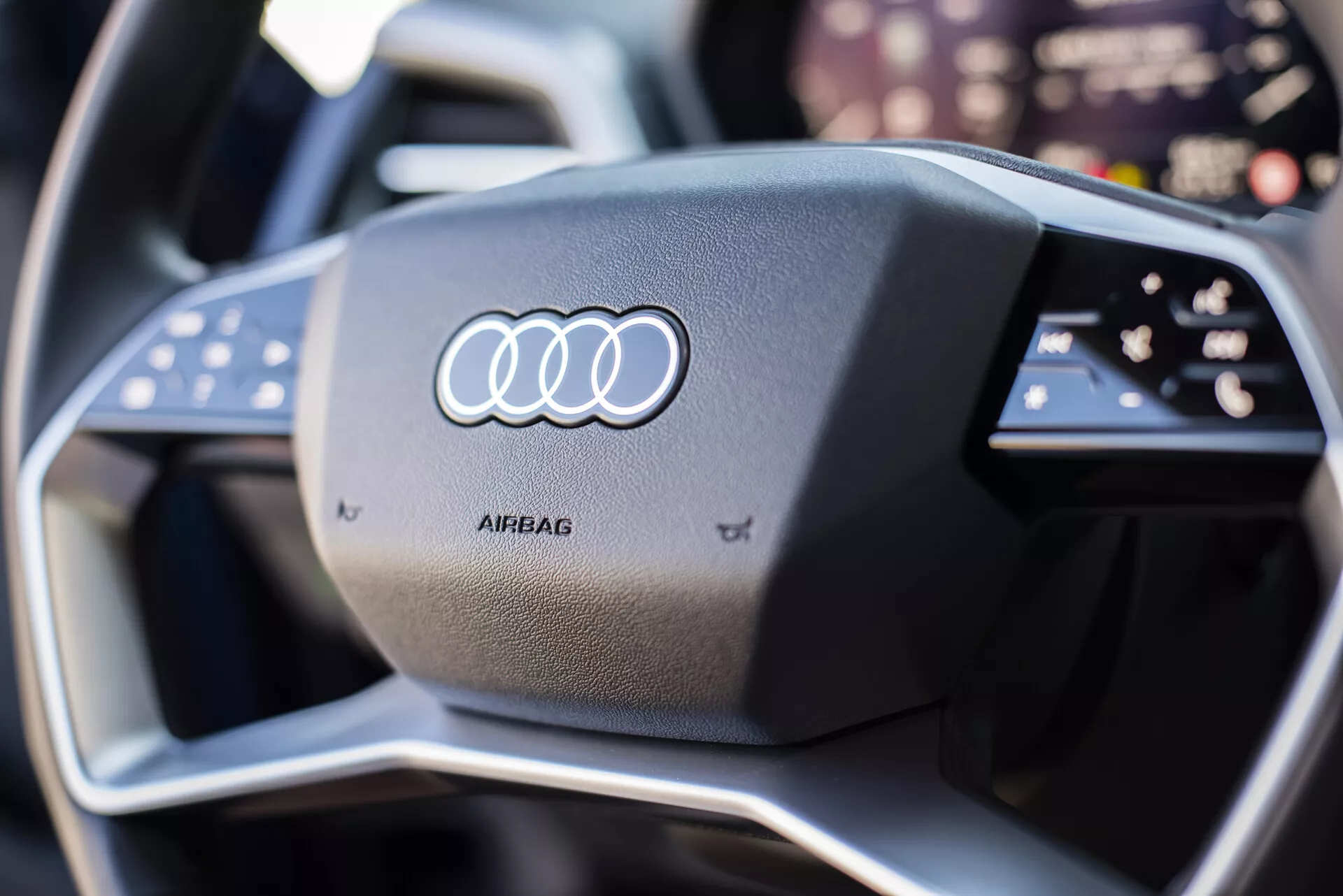 Detailný pohľad na volant Audi Q4 e-tron 2021 s ikonickým logom Audi na stredovej časti pre airbag, obklopený multifunkčnými tlačidlami, s moderným, športovým dizajnom vrátane kontrastného šitia a náznaku pokročilej technológie digitálneho prístrojového panela na pozadí