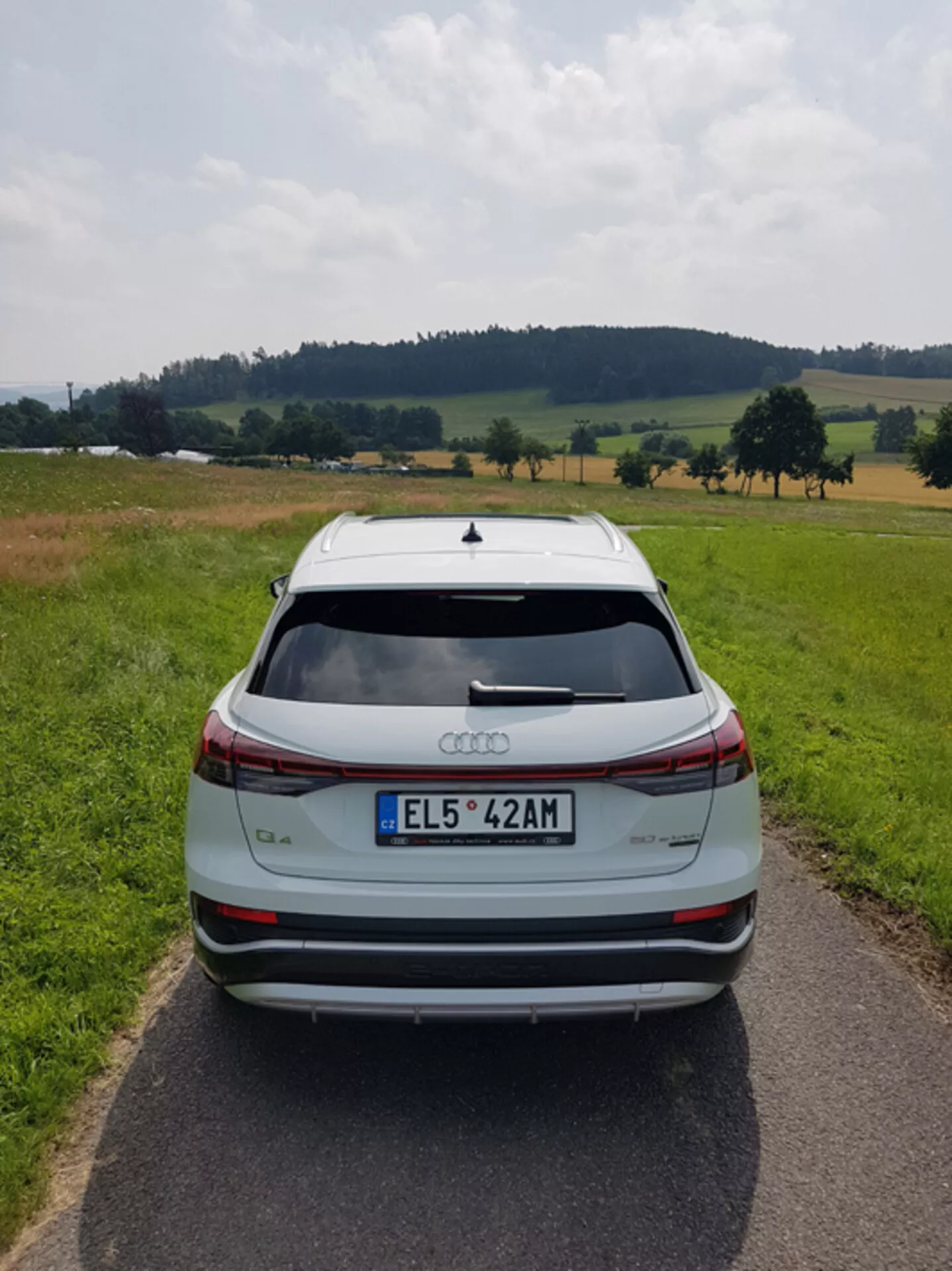 Zadný pohľad na strieborné Audi Q4 e-tron 2021 na úzkej spevnenej ceste uprostred pastierskej krajiny s poliami a stromami pod čiastočne zamračenou oblohou, zdôrazňujúci jeho štýlový dizajn, svetelný pruh brzdových svetiel, strešný spojler, senzory parkovania vzadu a dvojfarebné zliatinové kolesá.