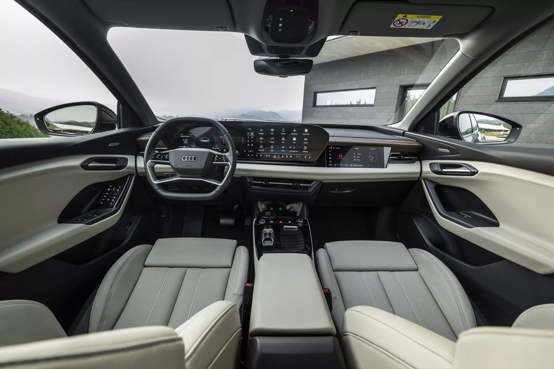 Pohľad na interiér Audi Q6 e-tron 2024 z perspektívy zadného sedadla so zameraním na predné sedadlá. Svetlé kožené predné sedadlá sú vybavené nastaviteľnými opierkami hlavy a elektronickými ovládačmi. Palubná doska zobrazuje široko rozsiahly digitálny prístrojový panel a veľkú centrálnu dotykovú obrazovku pre infotainment, obklopenú ovládacími tlačidlami pre klimatizáciu a jazdné režimy. Stredová konzola obsahuje držiaky na nápoje, úložný priestor a automatický radič prevodovky. Volant je vybavený niekoľkými ovládacími prvkami a panoramatická strecha naznačuje množstvo prirodzeného svetla. Celkovo obraz zdôrazňuje pokročilé technológie a luxusné automobilové prvky.