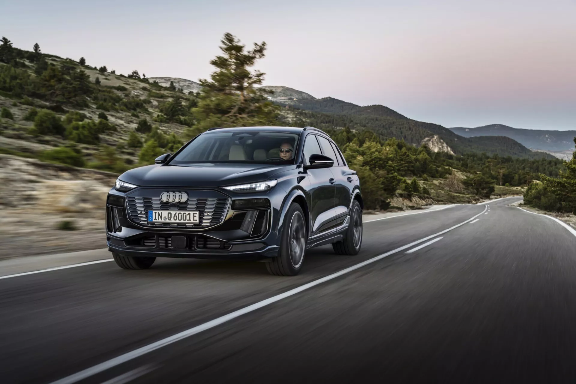 Audi Q6 e-tron 2024 s moderným dizajnom, veľkou prednou mriežkou, elegantnými LED svetlometmi a tvarovanými líniami karosérie. Pohľad zo trištvrte z predu, dynamický záber v pohybe na kopcovitej ceste, zdôrazňujúci výkon a ovládateľnosť.