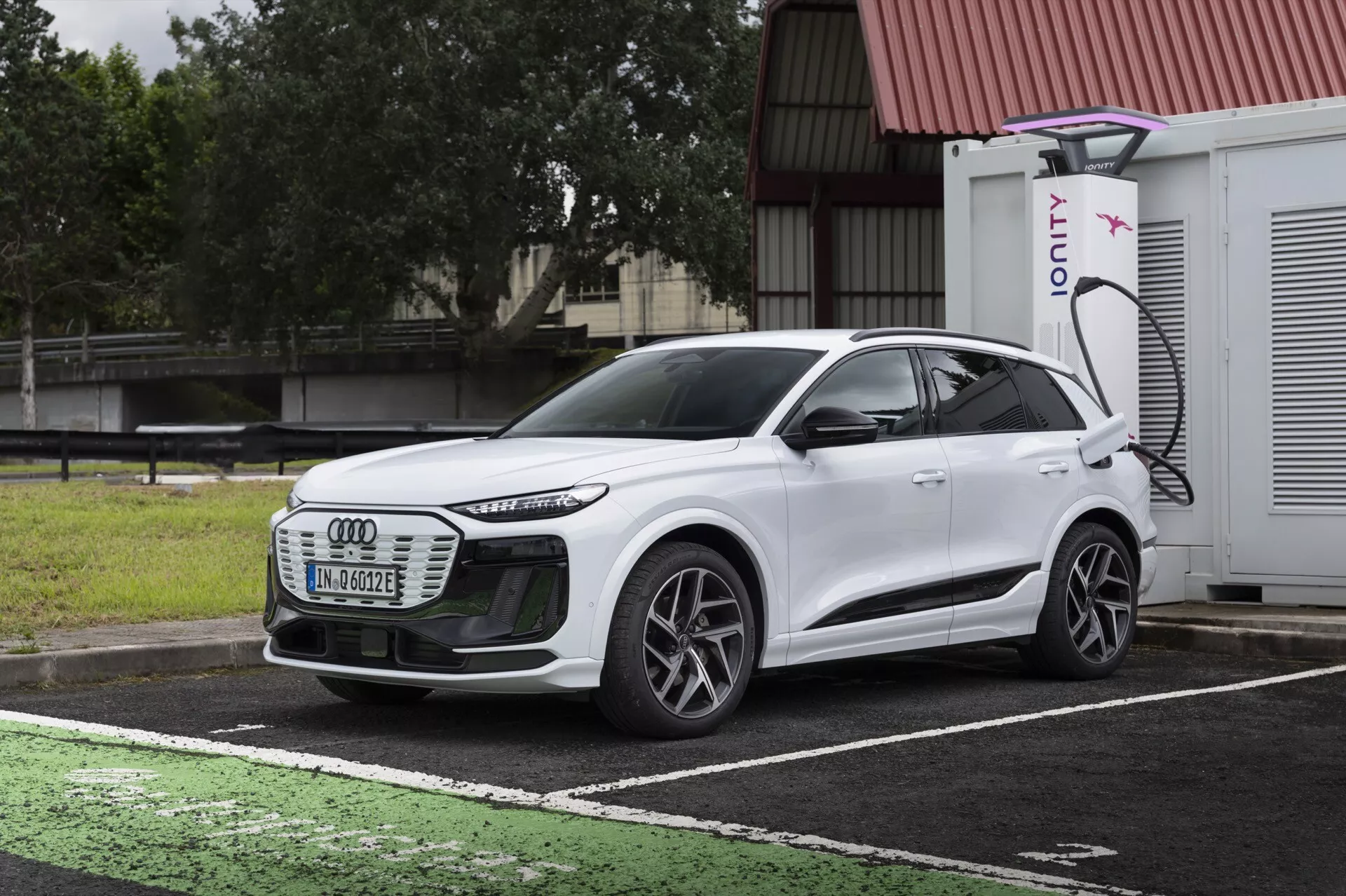 Trojštvrťový pohľad spredu na biely elektrický SUV Audi Q6 e-tron 2024, zvýrazňujúci prednú mriežku, LED svetlomety, aerodynamické línie, viacramenné kolesá a bočný profil. Vozidlo je zaparkované na vyhradenom mieste pre nabíjanie elektrických vozidiel so viditeľným nabíjacím portom a nabíjačkou IONITY v pozadí. Obrázok kontrastuje dizajn auta s tmavou, zamračenou oblohou a mokrým chodníkom, ilustrujúc modernú technológiu elektrických vozidiel a pokročilé osvetlenie.