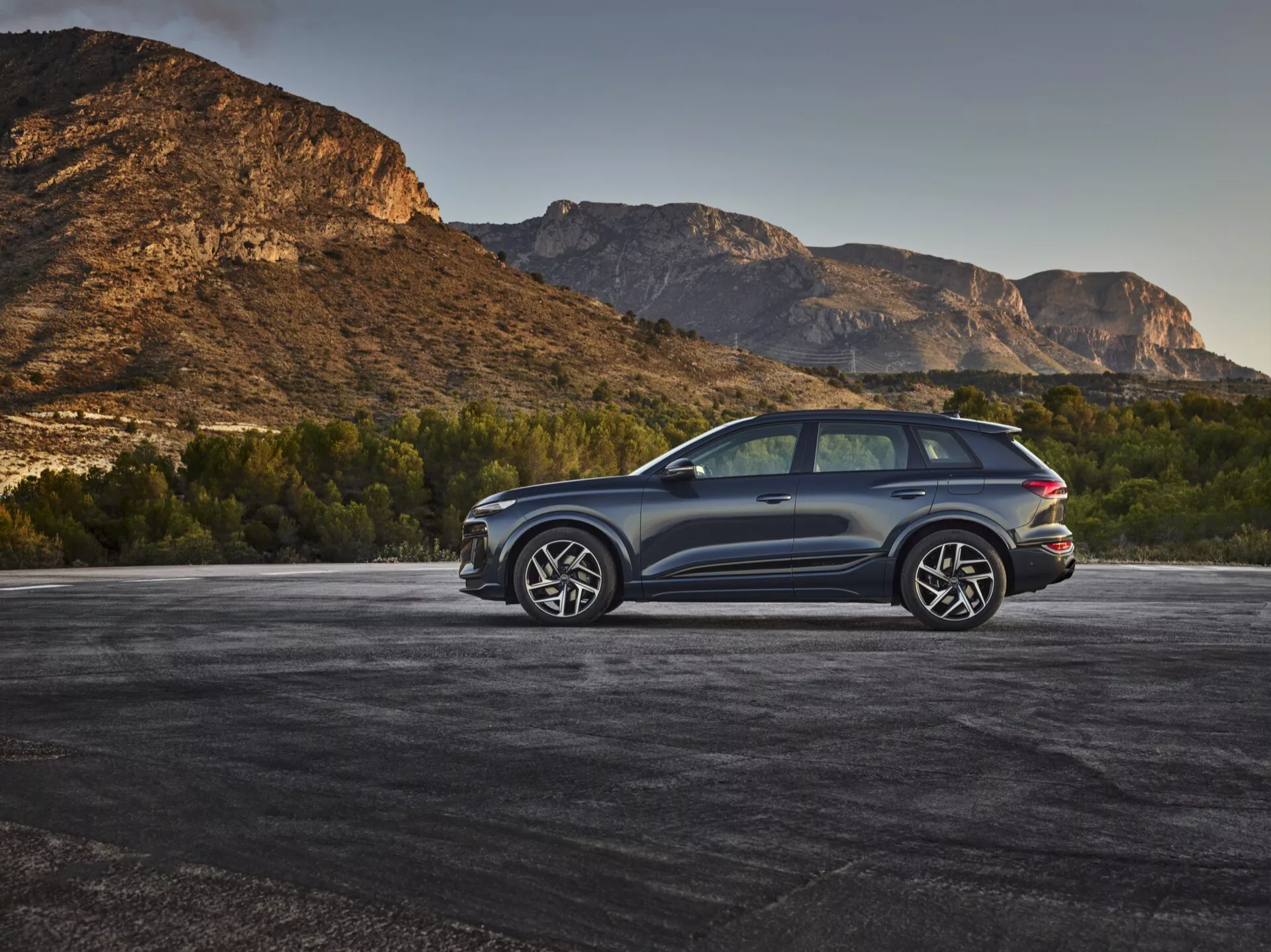 Moderné SUV, pravdepodobne Audi Q6 e-tron 2024, zaparkované vonku s horským pozadím, zachytené v troch štvrtinách zadného pohľadu z vyvýšeného uhla. Tmavá metalická farba karosérie kontrastuje s prírodným prostredím, čo zvýrazňuje elegantné línie a aerodynamické kontúry. Viditeľné detaily zahŕňajú LED zadné svetlá, veľké zliatinové kolesá s viaclúčovými dizajnmi, dvojitý výfukový systém, tónované okná, panoramatické strešné okno a ochranné lišty pozdĺž spodných hrán. Zadné okno je mierne šikmé so zadným spojlerom a veľkými bočnými zrkadlami pravdepodobne vybavenými integrovanými smerovkami, čo zvyšuje športový a luxusný vzhľad vozidla.