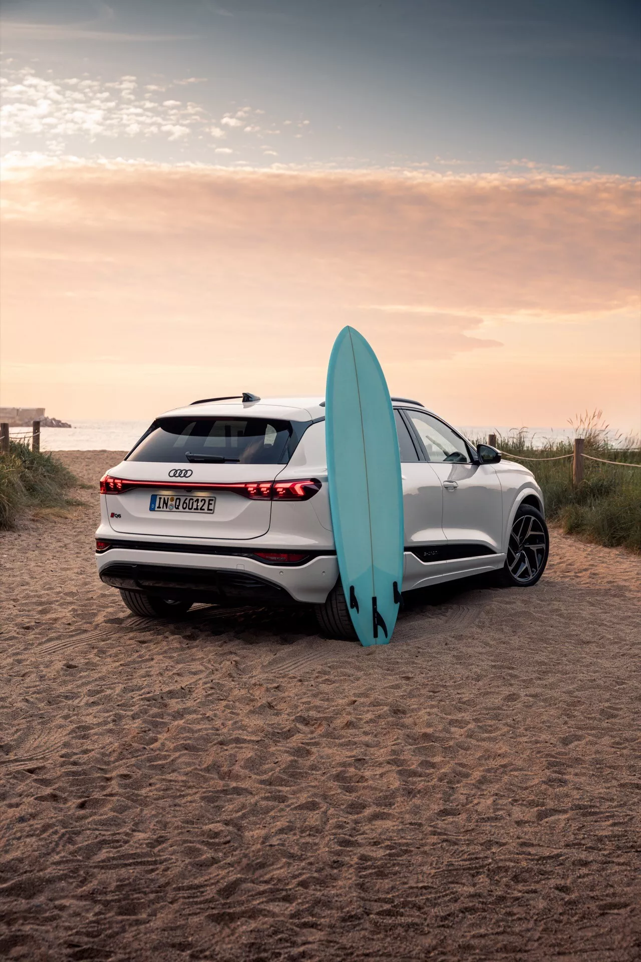 Obrázok zobrazuje biele SUV Audi Q6 e-tron 2024 zaparkované na piesočnatom pobreží s surfovacou doskou. Fotografované zozadu z trojštvrťového uhla zachytáva plynulé línie vozidla, LED zadné svetlá, parkovacie senzory, cúvaciu kameru a viaclúčové zliatinové kolesá. Odraz zapadajúceho slnka zvýrazňuje pokročilú kvalitu laku, zatiaľ čo surfová doska opretá o vozidlo naznačuje prvok dobrodružného životného štýlu.