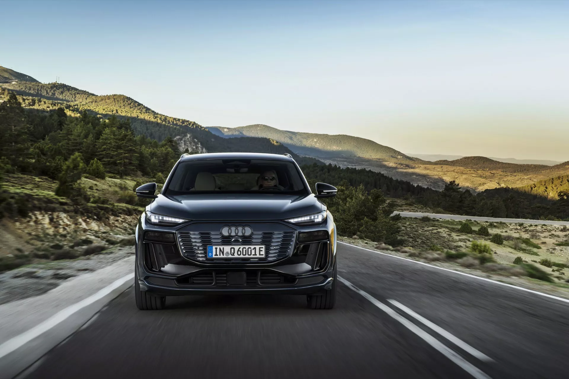 Čelný pohľad na Audi Q6 e-tron 2024, ktorý ukazuje prednú mriežku, svetlomety a nárazník so štyrmi vzájomne prepojenými kruhmi ako logom. Európska poznávacia značka je namontovaná na mriežke a označenie 'e-tron' naznačuje jeho elektrickú/hybridnú povahu. Auto jazdí po otvorenej ceste s horskou krajinou v pozadí, čo naznačuje nastavenie neskorého popoludnia. Funkcie zahŕňajú elegantné LED svetlomety, estetickú mriežku, senzory/kamery asistencie vodiča, aerodynamické zrkadlá a efektívny dizajn.