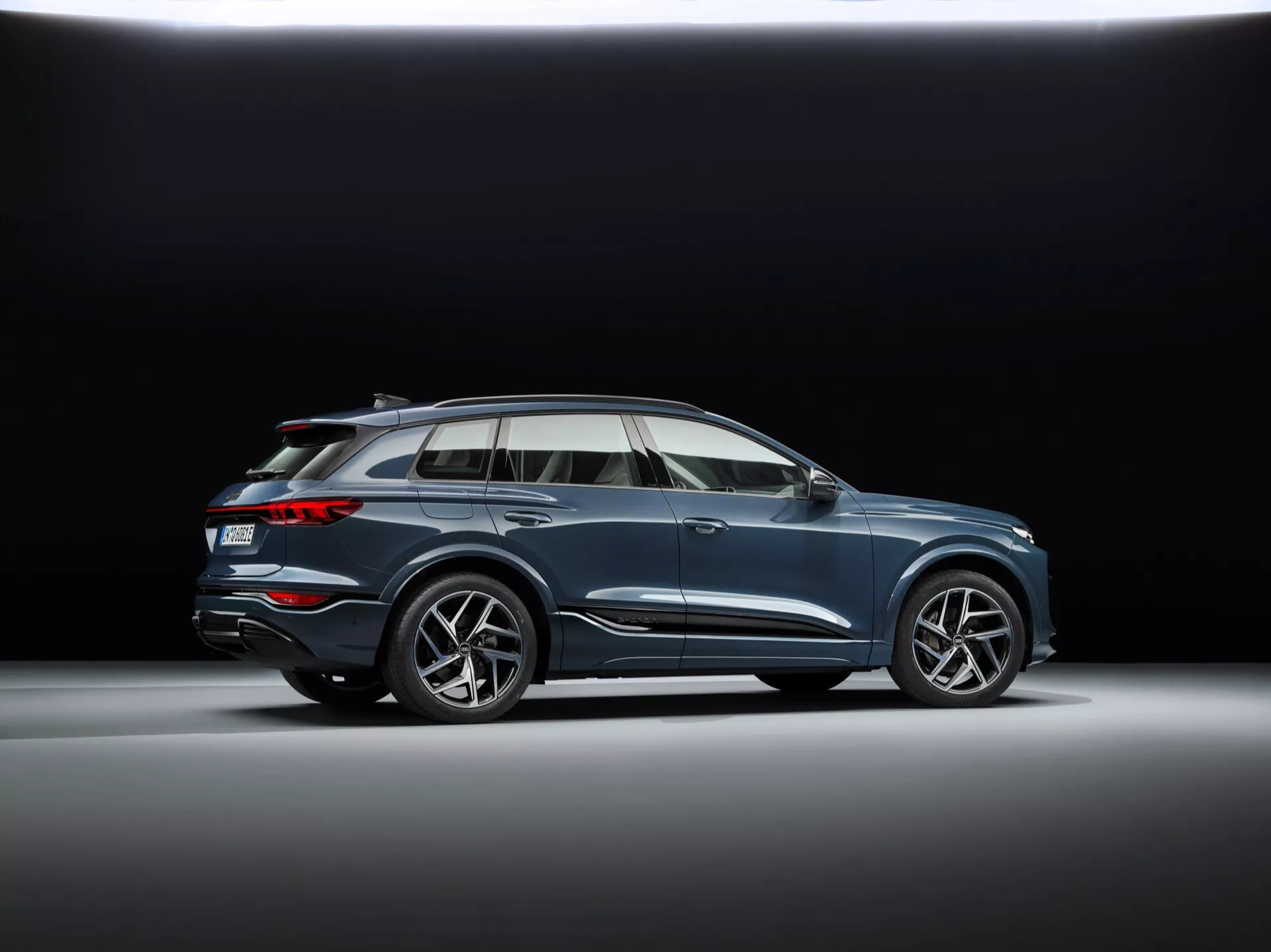 Nízkouhlový profilový záber Audi Q6 e-tron 2024 v štúdiovom nastavení s tmavým pozadím, so športovými viac lúčovými zliatinovými kolesami, plynulým dizajnom karosérie s charakterovou líniou, aerodynamickými spätnými zrkadlami s integrovanými smerovkami, chrómovanými okennými lištami, panoramatickým strešným oknom, strešnými lyžinami, mierne zvýšenou svetlou výškou a zapusteným dvojitým výfukovým systémom.