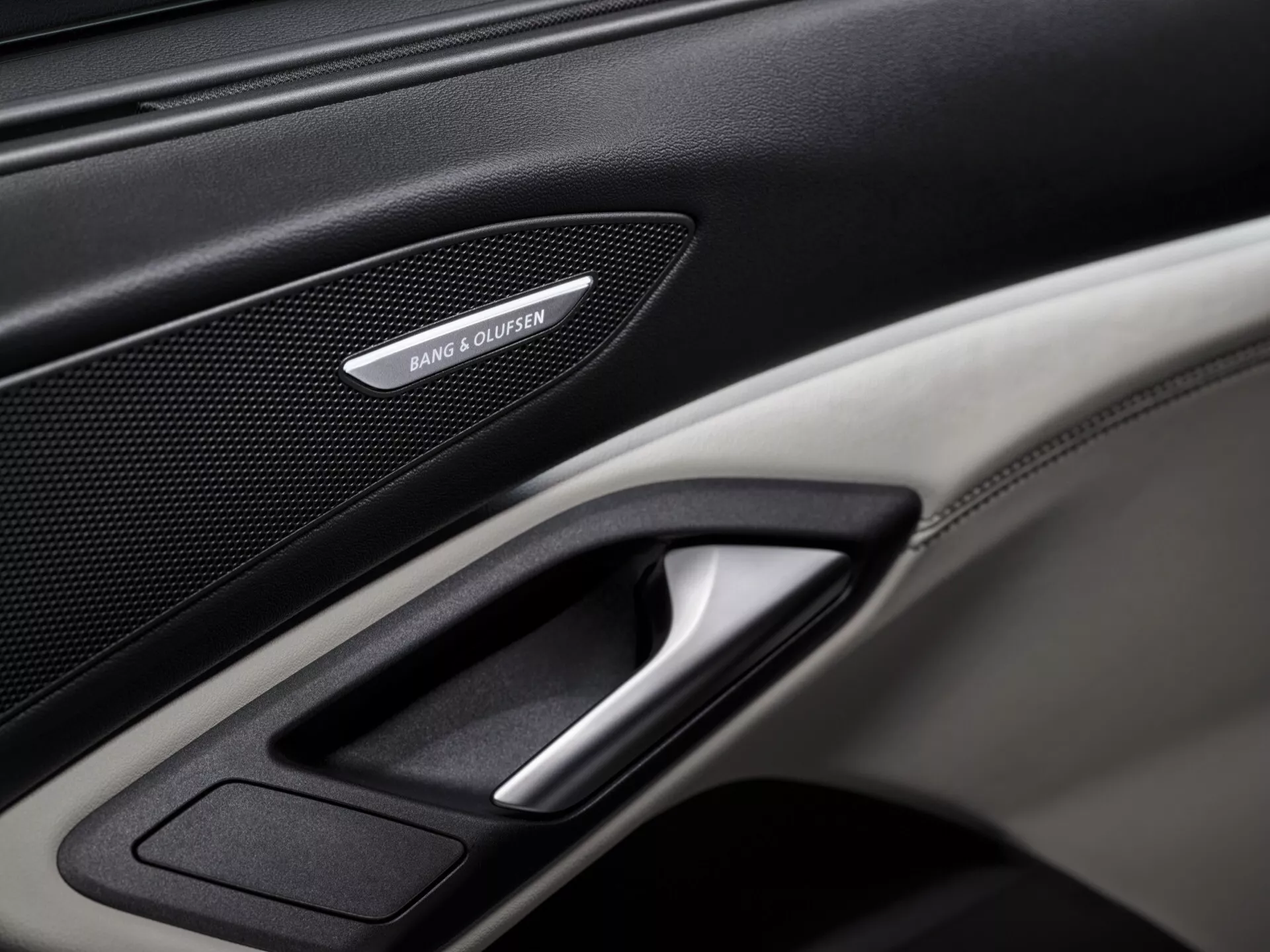 Detailný záber na interiérový dverový panel Audi Q6 e-tron 2024 GF, zdôrazňujúci prémiové prvky ako je mriežka reproduktora Bang & Olufsen, pochrómovaná páčka dverí a kombinácia kože a textúr z uhlíkových vlákien. Dobre osvetlený obrázok sa zameriava na detailné prvky panelu, predstavujúce luxusné a technologické prvky vozidla.