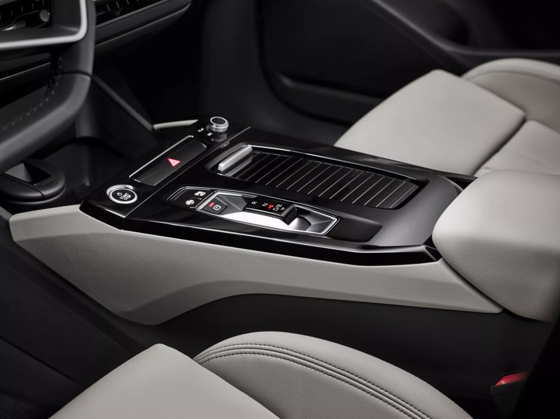 Záber interiéru Audi Q6 e-Tron 2024 zameraný na oblasť stredovej konzoly. Obrázok, urobený z pohľadu spolujazdca, zvýrazňuje lesklú čiernu radiacu páku s kovovými detailmi, niekoľko ovládacích tlačidiel, malý úložný priestor s posuvným krytom, dva držiaky na poháre, kožou čalúnenú lakťovú opierku, vetracie otvory klimatizácie a časti svetlo zafarbených kožených sedadiel s kontrastnými tmavšími sekciami. Funkcie stredovej konzoly ukazujú moderný dizajn a luxusné prvky.