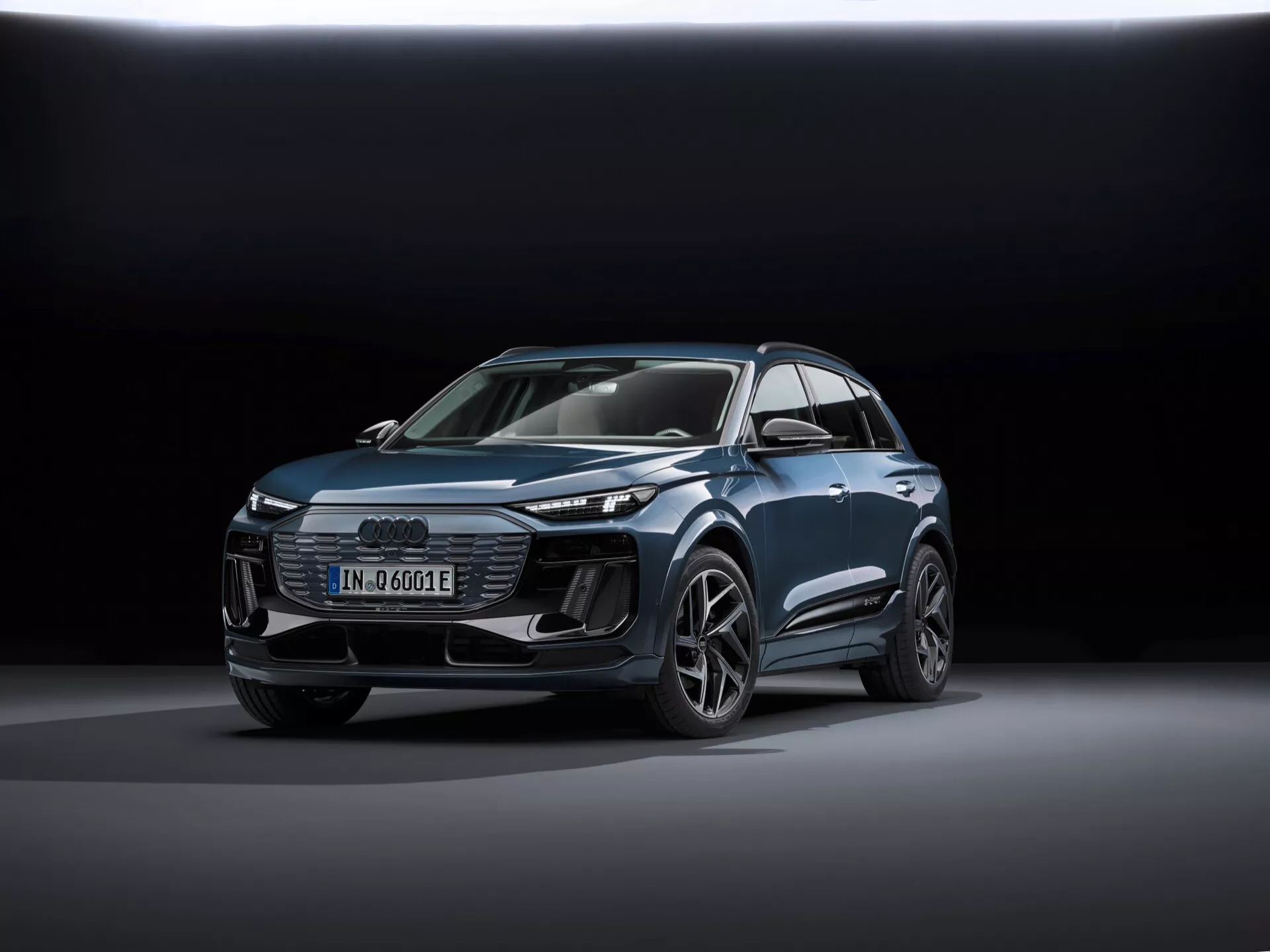 Obrázok zobrazuje moderné SUV Audi Q6-e-tron 2024 v pohľade z troch štvrtín spredu, zvýrazňujúc predný aj bočný profil. Medzi hlavné identifikátory patrí znak štyroch kruhov Audi na mriežke a znak 'e-tron', ktorý naznačuje elektrický model. Záber je zhotovený z nízkej perspektívy, čo zvýrazňuje siluetu vozidla a ukazuje jeho dizajnové línie, svalnaté oblúky kolies a aerodynamické prvky. Auto je umiestnené v strede rámca, stojí na hladkom reflexnom povrchu s mäkkým, rozptýleným osvetlením a tmavým prechodovým pozadím, čo zdôrazňuje dizajn vozidla. Medzi viditeľné technologické prvky patria LED svetlomety, denné svetlá (DRL), digitálny displej a zložito navrhnuté zliatinové kolesá. Celková leštená prezentácia naznačuje propagačnú fotografiu, ktorá zvýrazňuje technológiu, dizajn a luxusné aspekty vozidla.