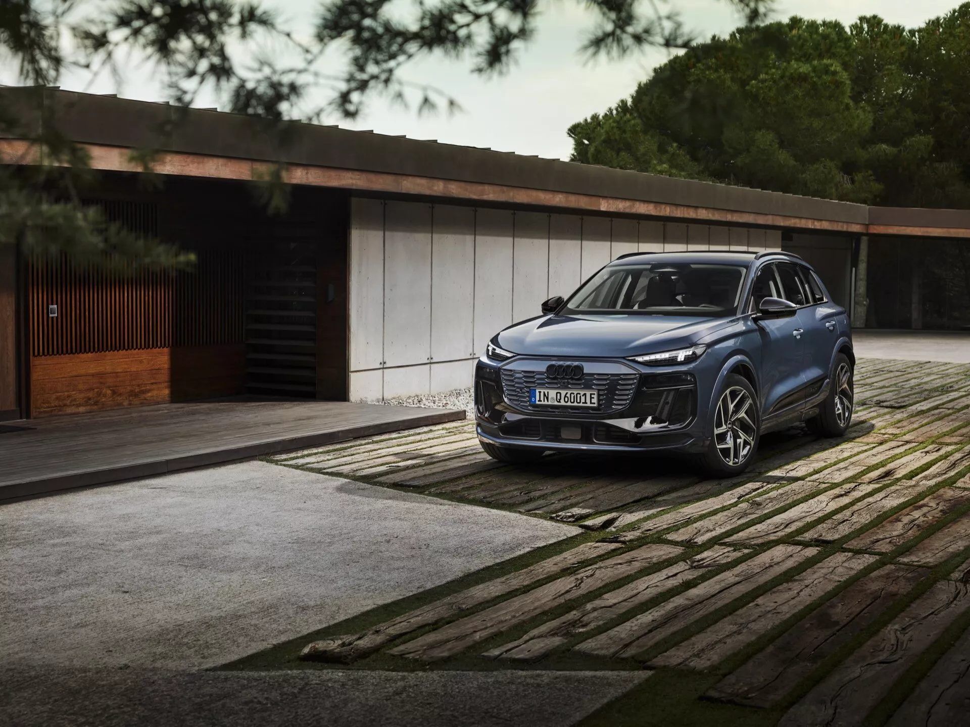 2024 Audi Q6 E-Tron trojštvrťový predný pohľad SUV s elegantnými LED svetlometmi, výraznou mriežkou a veľkým emblémom zaparkovaným na vzorovanej dlažbe. Luxusná budova a stromy v pozadí zdôrazňujú luxus a exkluzivitu.