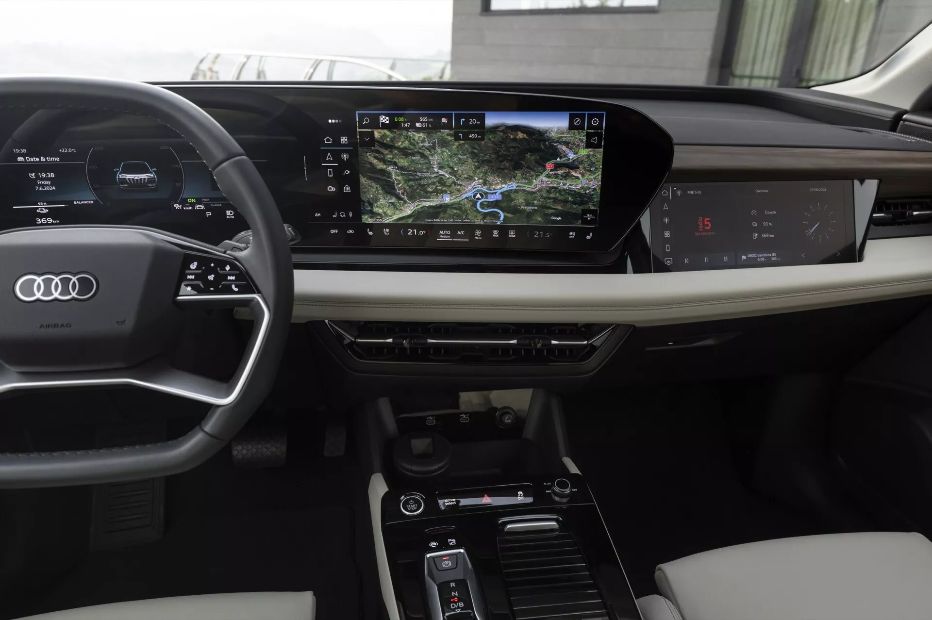 Interiér moderného Audi Q6 e-tron 2024, zobrazený z pohľadu vodiča. Vybavenie zahŕňa kožený volant s tromi ramenami so zabudovanými ovládacími prvkami, digitálny prístrojový panel zobrazujúci rýchlosť a navigáciu a horizontálne orientovaný dotykový infotainment systém. Stredová konzola obsahuje ovládanie klimatizácie a jazdných režimov. Luxusné materiály ako koža a drevené obloženie vylepšujú estetiku.