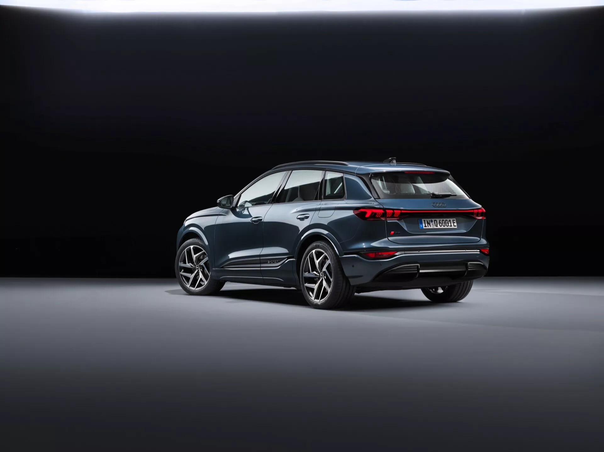 Audi Q6 e-tron 2024 (gf) je zobrazené v zadnom trojštvrťovom pohľade, ukazujúc moderné SUV diagonálne umiestnené na hladkej, reflexnej podlahe showroome. Pozadie sa mení z tmavého na svetlé, čím zvýrazňuje vozidlo. Viditeľné prvky zahŕňajú prepojené zadné svetlá, výrazný strešný spojler, výrazné oblúky kolies s veľkými zliatinovými kolesami, chrómovanú lištu pozdĺž okien, zadný nárazník s reflektormi a senzormi a dvojitý výfukový systém, ktorý naznačuje výkon, čo môže znamenať, že ide o hybridný alebo elektrický model.