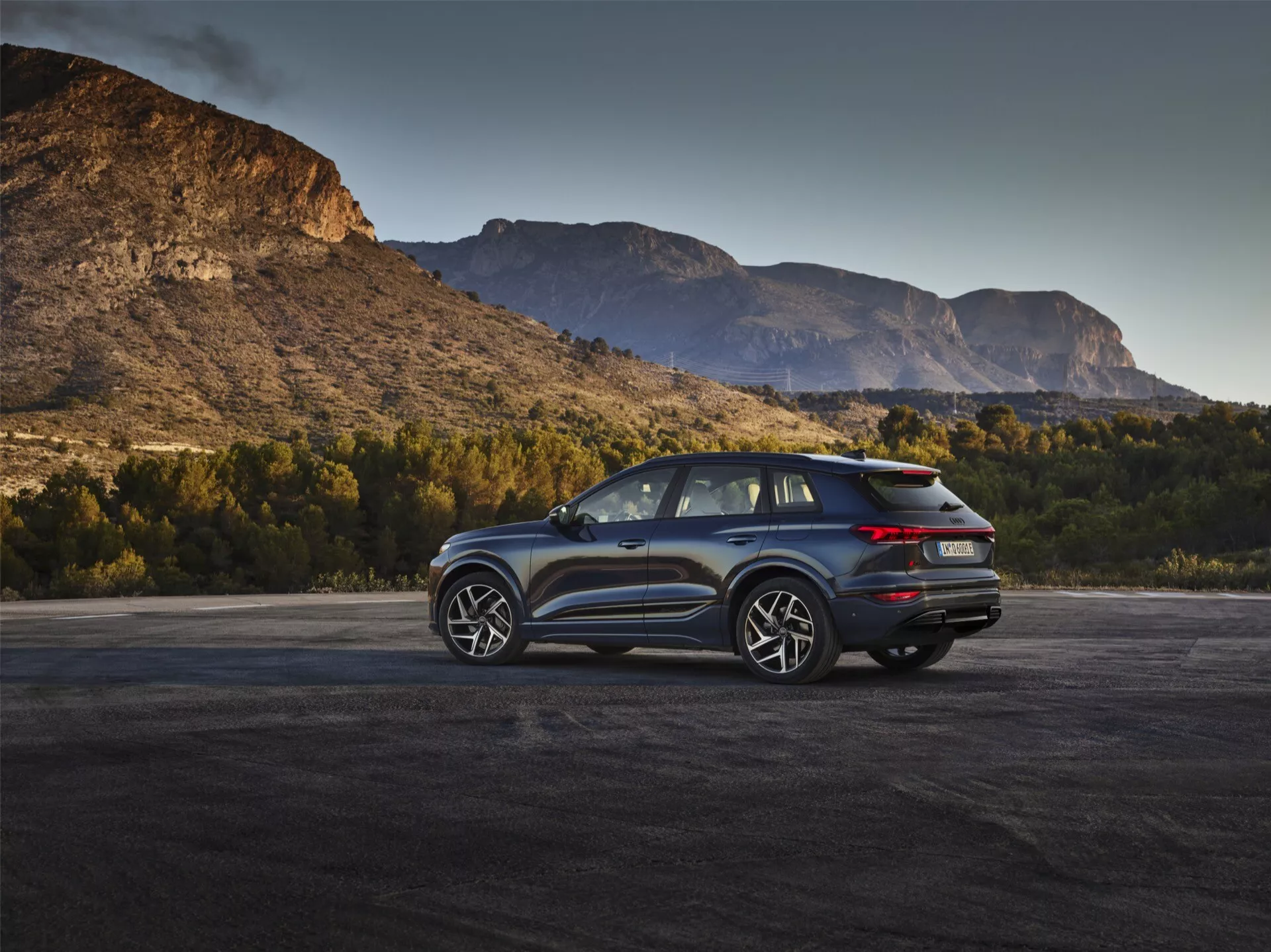 Obrázok ukazuje moderné SUV Audi Q6 e-Tron 2024 v vonkajšom prostredí s malebným horským pozadím počas svitania alebo súmraku. Trištvrte zadný uhol zvýrazňuje výrazné LED zadné svetlá, čistý dizajn zadného veka s možnou zadnou kamerou a dvojitý výfukový systém. Bočný profil zobrazuje viacpaprskové zliatinové kolesá, veľké okná a sklonenú líniu strechy, čo naznačuje športovú alebo luxusnú výbavu s možnými aerodynamickými funkciami. Zrkadlá môžu integrovať technológiu monitorovania slepého uhla. Prirodzené osvetlenie zvyšuje príťažlivosť vozidla, zvyšuje dojem dobrodružstva a schopností.