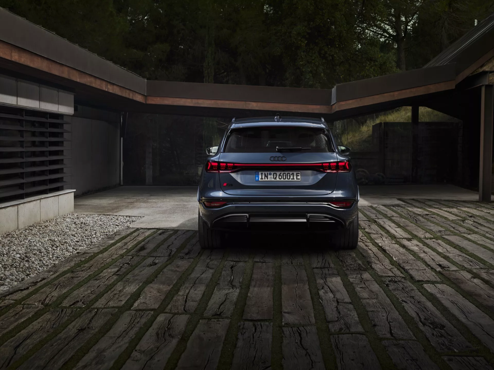 Zadný uhol pohľadu na Audi Q6 e-tron 2024, ktorý zdôrazňuje jeho čistý, elegantný a moderný dizajn. Viditeľné kľúčové prvky zahŕňajú súvislý svetelný pruh prepojujúci obe zadné svetlá, označenie modelu a elektrického pohonného systému, integrovaný zadný nárazník so športovým difúzorom, dopredu naklonené zadné sklo naznačujúce SUV alebo crossover, skryté výfukové koncovky a stredne umiestnenú registračnú značku. Auto je zaparkované na dláždenom príjazdovom ceste s náznakom moderného garáže v pozadí, naznačujúc luxusný a ekologický životný štýl.