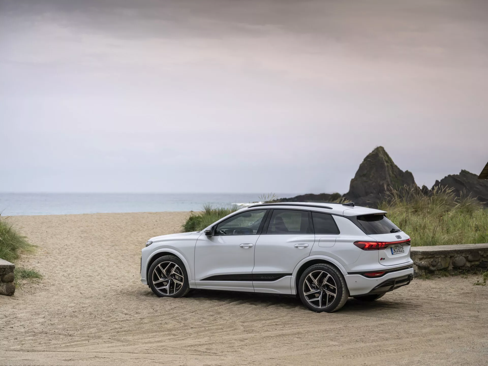 Biele SUV Audi Q6 e-tron 2024 je zaparkované na prašnej ceste s plážovým pozadím, zachytené z trojštvrťového zadného pohľadu. Obrázok zdôrazňuje moderné dizajnové prvky vozidla vrátane zadných svetiel, zadného nárazníka, štýlových kolies a elegantných línie. Vyššia svetlá výška a celkový postoj naznačujú jeho vhodnosť pre rôzne terény. Mäkké rovnomerné osvetlenie naznačuje zamračený deň alebo blízky východ/západ slnka, čím zvýrazňuje dobrodružný charakter. Viditeľné sú moderné zliatinové kolesá, zadný spoiler a LED zadné svetlá, čo signalizuje pokročilú technológiu a športový charakter.