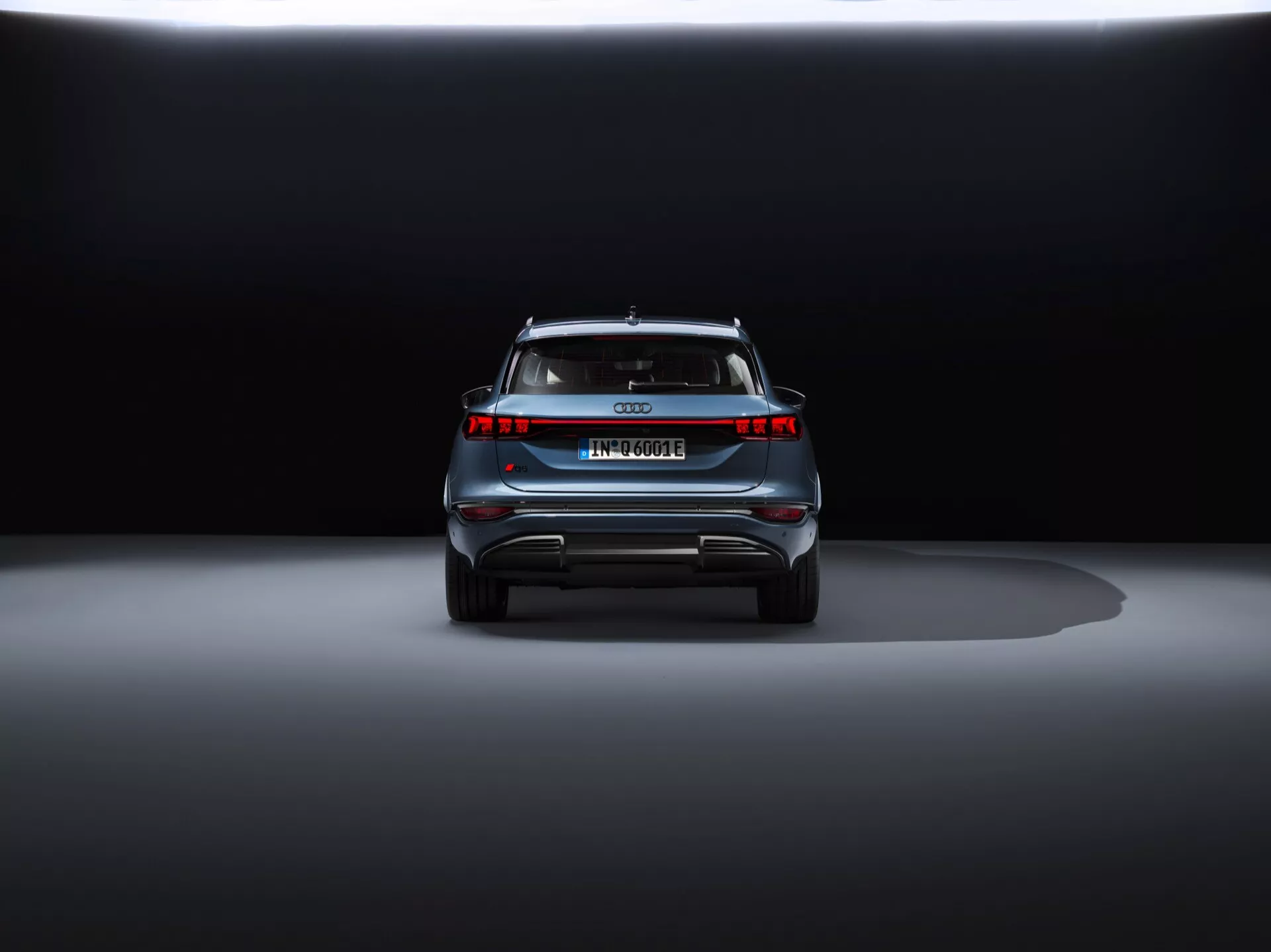 Zadný pohľad na Audi Q6 e-tron 2024 na výstave s dramatickým osvetlením. Snímek na úrovni zeme zdôrazňuje moderné zadné LED svetlá, absenciu výfukových trubiek, celoplošné osvetlenie, európsku poznávaciu značku, strešný spojler a reflexné bezpečnostné prvky, čo naznačuje, že ide o luxusný elektromobil alebo hybrid