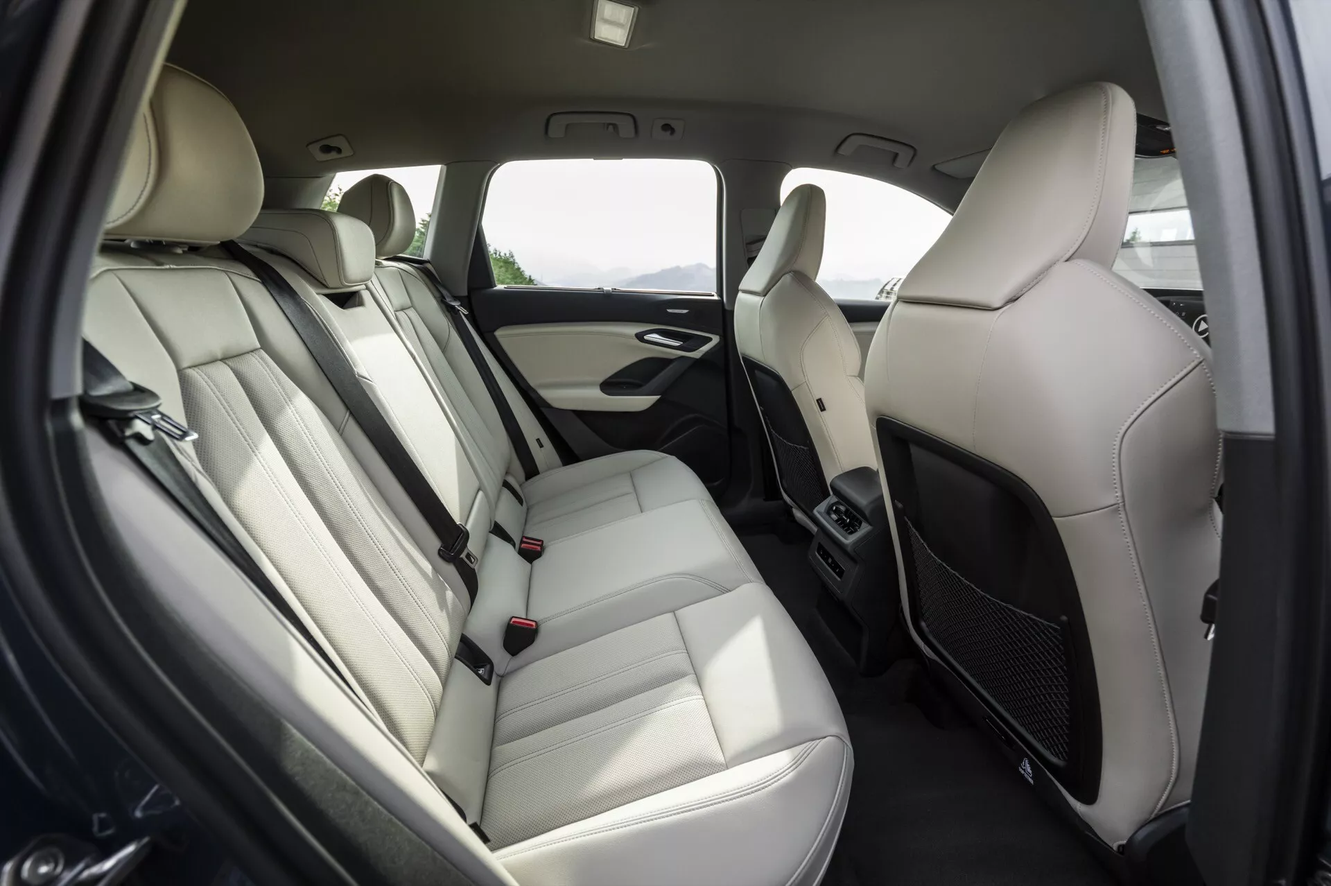 Interiér zadného priestoru pre cestujúcich modelu Audi Q6 e-tron 2024, so zameraním na usporiadanie zadných sedadiel. Lavicové sedadlá sú potiahnuté svetlou kožou s opierkami hlavy, opierkou a viditeľnými bezpečnostnými pásmi. Panel dverí obsahuje mriežku reproduktora, kľučku a pravdepodobne ovládanie okien. Farebná schéma je dvojfarebná, so svetlými a tmavými prvkami. Medzi prednými sedadlami sú ventilačné otvory klimatizácie, čo naznačuje klimatizáciu pre zadných pasažierov. Moderný, luxusný a pohodlný dizajn pre cestujúcich.