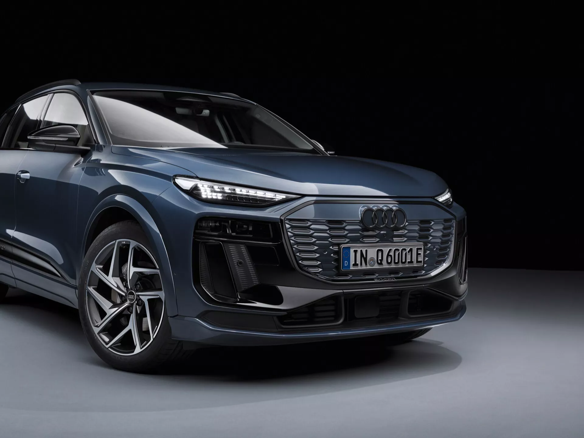 Obrázok zobrazuje moderné Audi Q6 e-tron 2024 v štúdiovom prostredí, zobrazené z predného trojštvrťového pohľadu, ktorý poskytuje jasný dojem z dizajnu vozidla zpredu a zboku. Jasne je viditeľný ľavý predný roh auta, vrátane prednej mriežky so vzorom medového plástu a centrálne umiestneným emblémom Audi, zložitých predných LED svetiel, ďalších vzduchových nasávacích otvorov alebo senzorov pod svetlami, športového predného nárazníka, veľkého hliníkového kolesa s nízkoprofilovou pneumatikou a obrysy bočného zrkadla. Obrázok zdôrazňuje elektrifikovaný modelový znak ('e') a zvýrazňuje jeho dizajnové a technologické prvky. Pozadie je jednoduché a tmavé, aby neodvádzalo pozornosť od samotného vozidla.