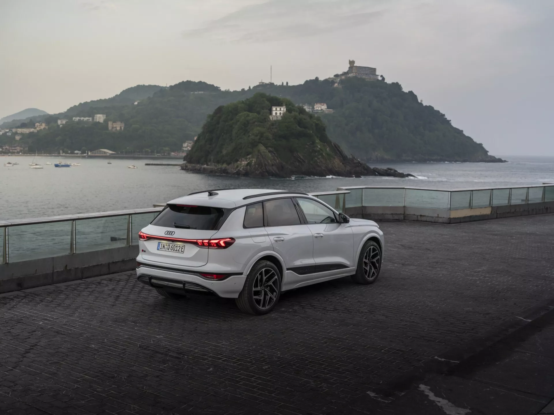 Trojštvrťový zadný pohľad na moderné, biele SUV Audi Q6 e-tron 2024 zaparkované na nábreží za súmraku, ukazujúce horizontálne zadné LED svetlá, znak výrobcu, zadný stierač, päťlúčové zliatinové kolesá, integrované smerové svetlá v zrkadlách, strešné nosiče a vytvarované línie karosérie na pokojným pozadím s malým ostrovom a budovou, zdôrazňujúc zmes úžitkových vlastností a výkonu.