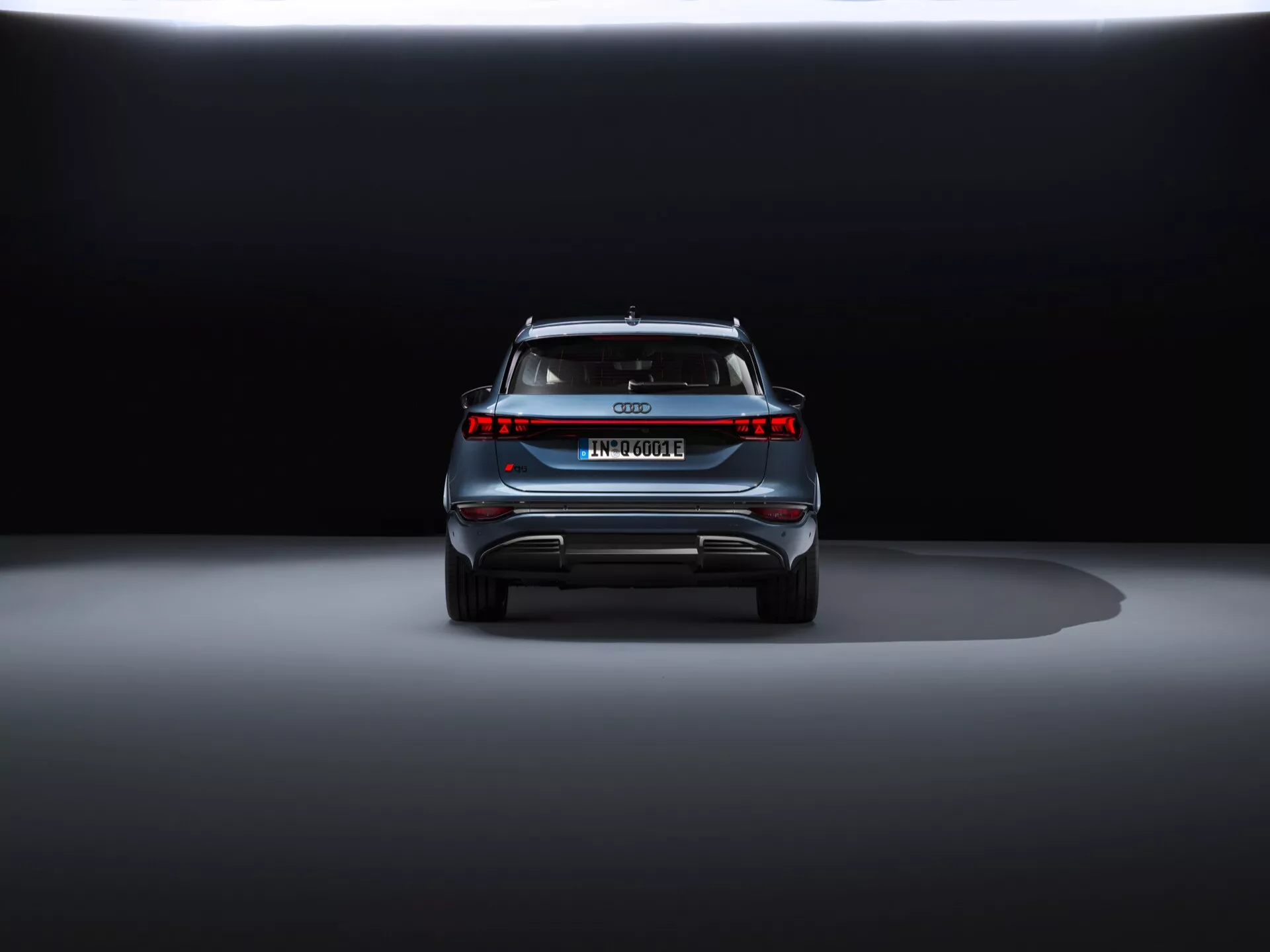 Zadný pohľad na Audi Q6 e-tron 2024 na tmavom pozadí, zobrazujúci štýlový dizajn, centrálny emblém, názov modelu, horizontálnu svetelnú lištu, zadný spojler a difúzorovitý nárazník s reflexnými prvkami.