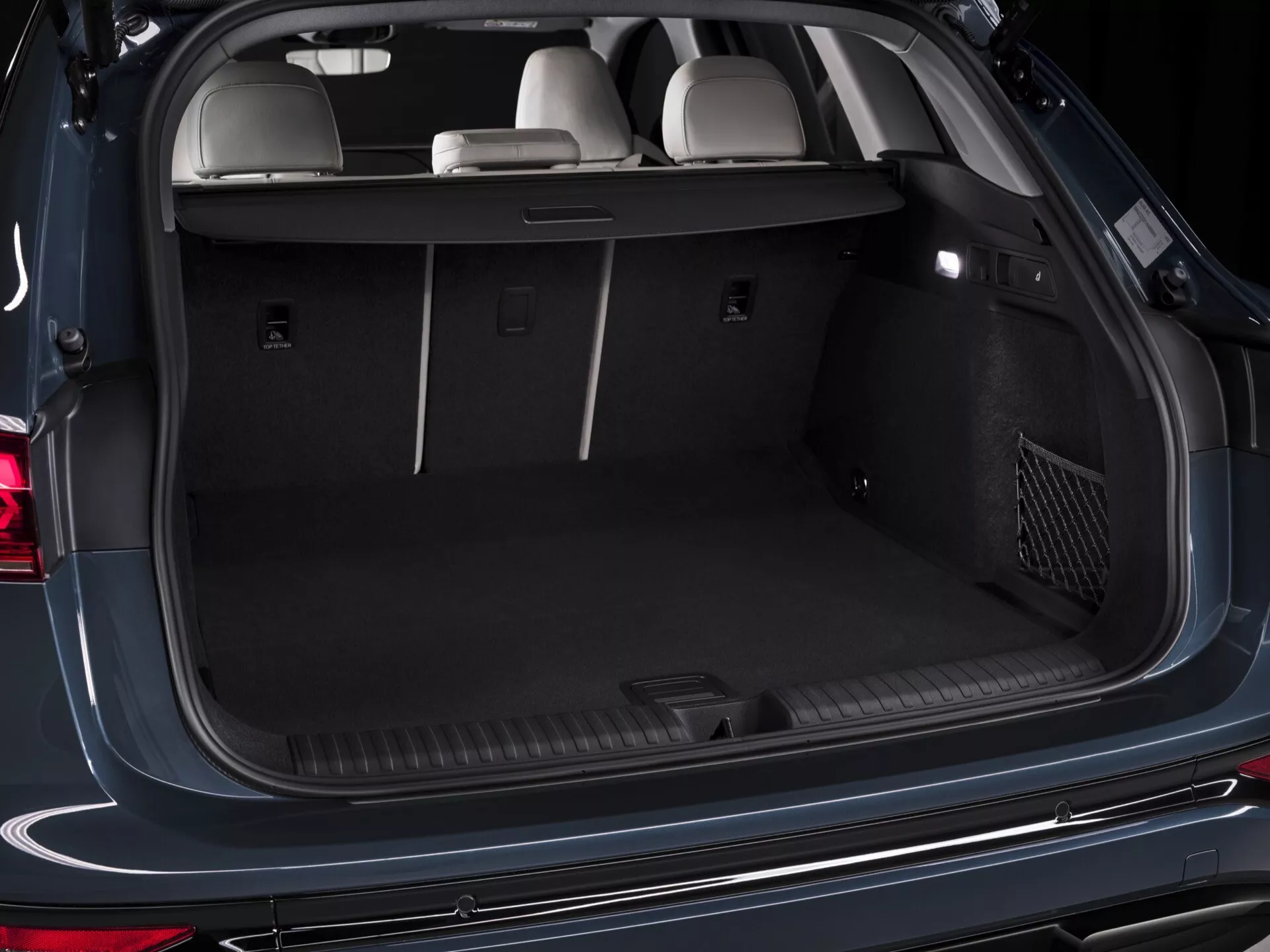 Detailný pohľad na otvorený batožinový priestor modelu Audi Q6 e-tron 2024 z pohľadu zozadu, ktorý zobrazuje jeho priestranný nákladný priestor. Funkcie zahŕňajú upevňovacie body, 12-voltovú zásuvku, zásuvný kryt batožinového priestoru, bočné úložné priehradky, vnútorné osvetlenie a sieťové vrecko. Interiér je dokončený tmavým kobercom a zadné sedadlá sú vo vzpriamenej polohe, čo zvýrazňuje kapacitu a variabilitu batožinového priestoru.