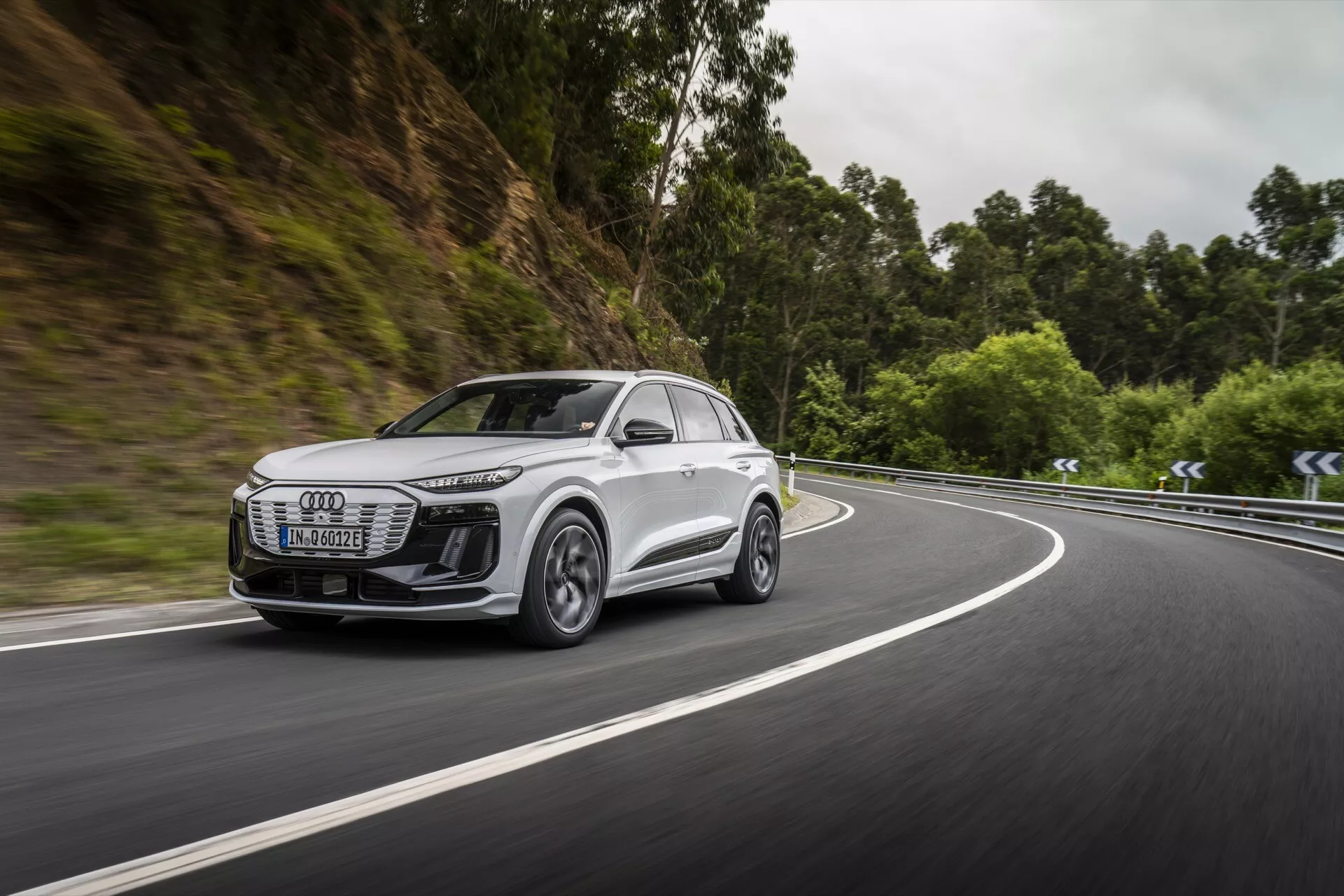 Obrázok zachytáva dynamický bočný predný pohľad na moderné biele SUV, pravdepodobne model Audi Q6 e-tron 2024, ktorý jazdí po ceste obklopenej lesným územím. Pozornosť je zameraná na prednú masku, svetlomety, bočný profil, kolesá a aerodynamiku bočných zrkadiel vozidla. Svetlomety sú elegantné s LED technológiou, čo naznačuje pokročilé osvetlenie. Kolesá zo zliatiny s viac paprskovým dizajnom naznačujú vysokovýkonné schopnosti. Pohyb vozidla na zákrute poukazuje na jeho jazdné vlastnosti a možné pokročilé systémy asistencie pri jazde. Rozmazané pozadie zdôrazňuje rýchlosť a efektívnosť v navigácii v náročných situáciách.