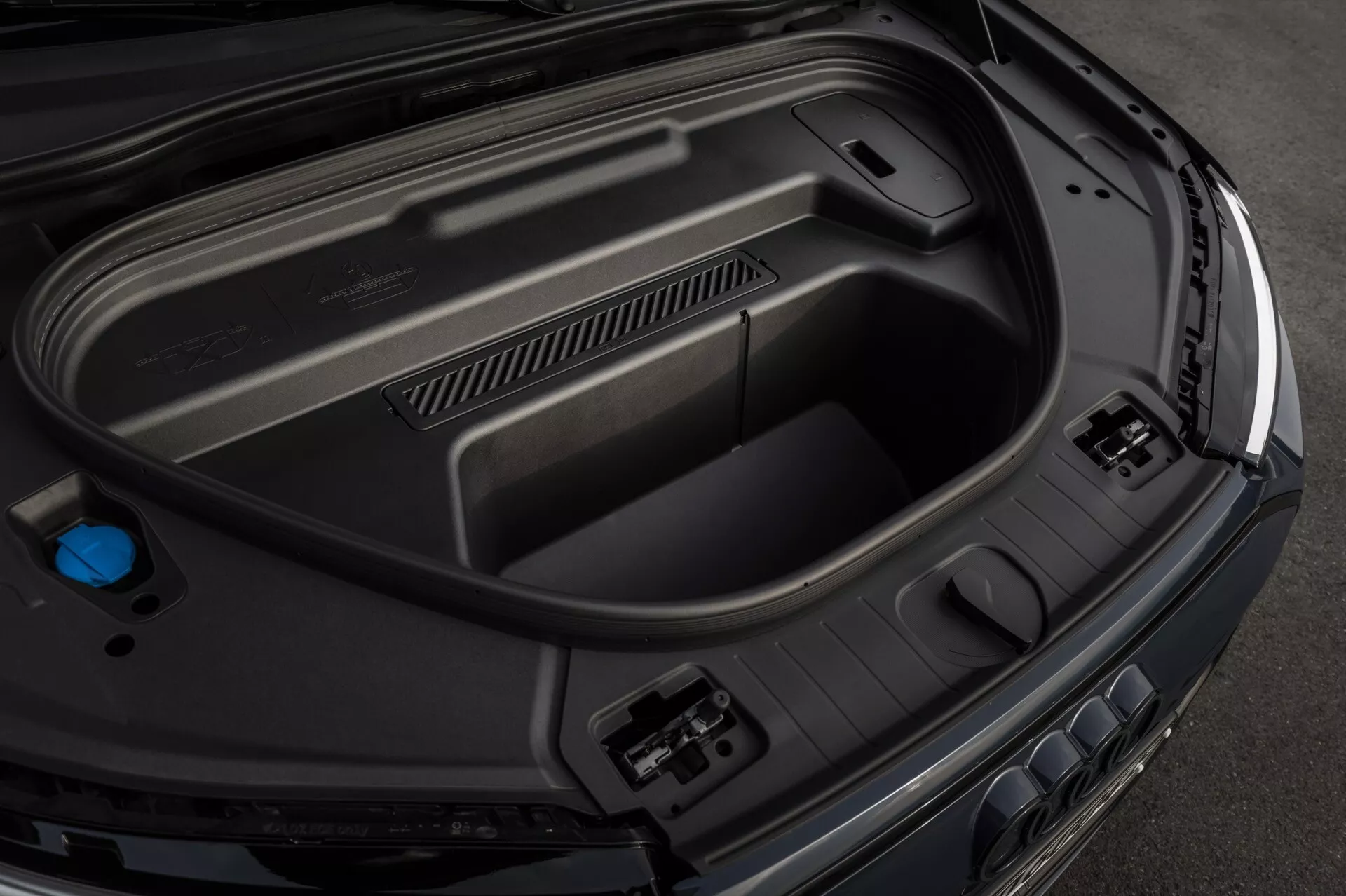 Detailný záber z vysokého uhla na zadný batožinový priestor Audi Q6-e-Tron 2024, ktorý zobrazuje vnútorné prvky vrátane tvarovaných plastových panelov pre odolnosť, zásuviek na príslušenstvo na pravej strane, zapusteného úložného priestoru na dne a modrého viečka na ľavej strane, čo naznačuje pokročilú automobilovú technológiu a elegantný, prémiový povrch.