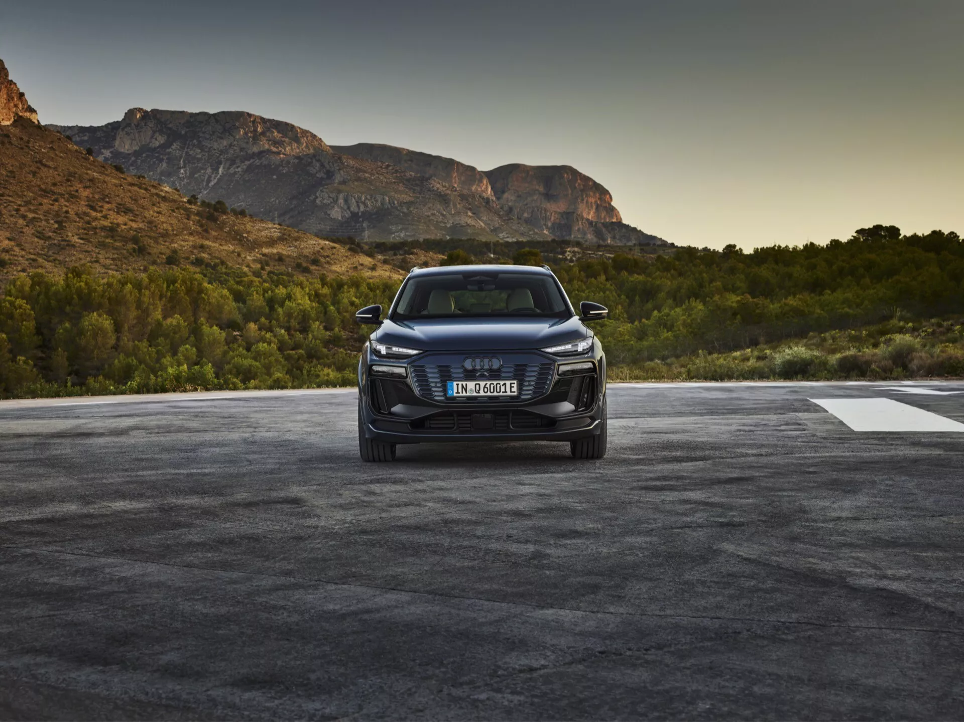 Obrázok vozidla Audi Q6 e-tron 2024 fotografovaného z predného trojštvrťového uhla, ktorý zobrazuje prednú mriežku vozidla s emblémom značky, ostré LED alebo laserové svetlomety a detailný predný nárazník s nasávacími otvormi a možnými senzormi alebo kamerami naznačujúcimi asistenčné systémy pre vodiča. Kapota obsahuje kontúrové línie a elegantný bočný profil naznačuje aerodynamiku s vyvýšeným postojom SUV. Vozidlo je umiestnené v exteriéri s prírodnou scenériou, ktorá naznačuje východ alebo západ slnka.